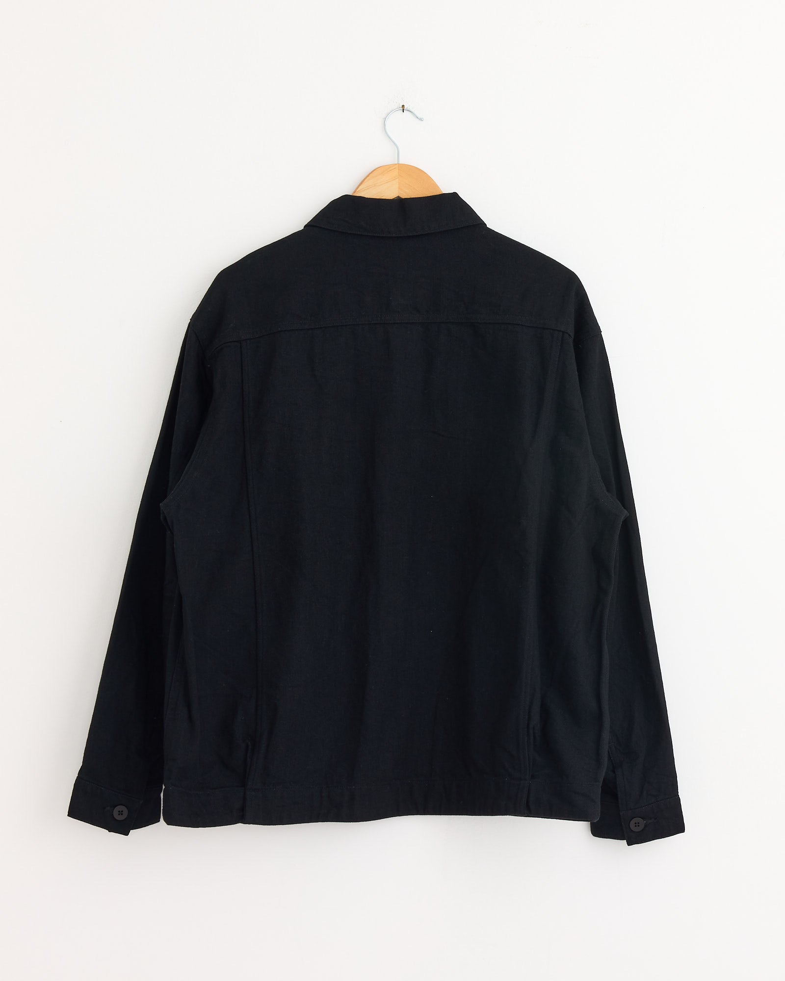 The Still By Hand 12oz Denim Jacket in Black, made in Japan, hangs on a wooden hanger against a white wall, displayed from the back to show its long sleeves and collar.