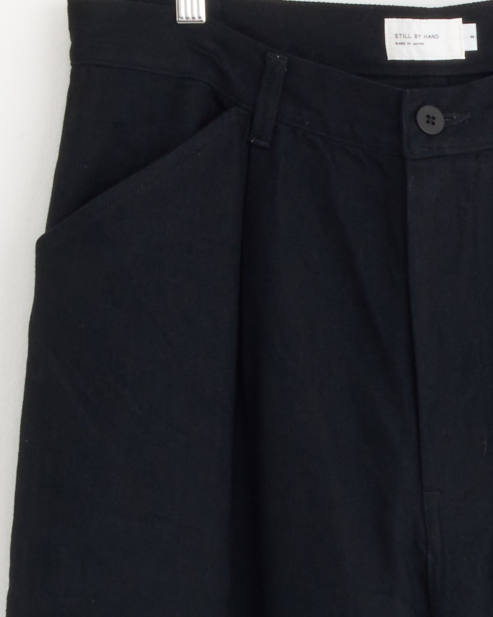 Close-up of the Still By Hand 12oz Denim 1 Tuck Pant in Black, featuring a button, zipper, side pocket, and a white inner waist label. Made in Japan from 12oz denim.