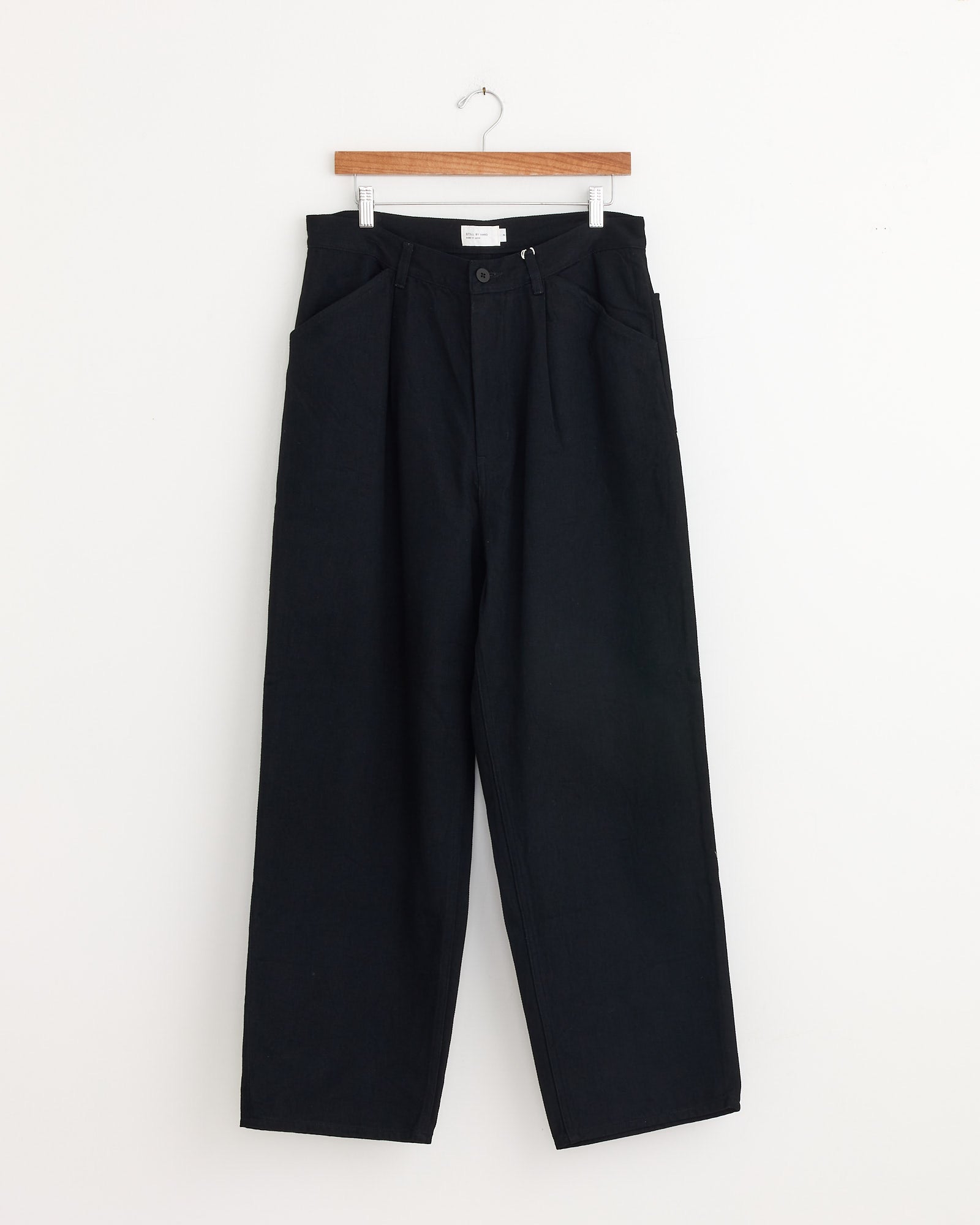 The Still By Hand 12oz Denim 1 Tuck Pant in Black, featuring a high-waisted, wide-leg design, hangs on a wooden hanger against a plain white wall.