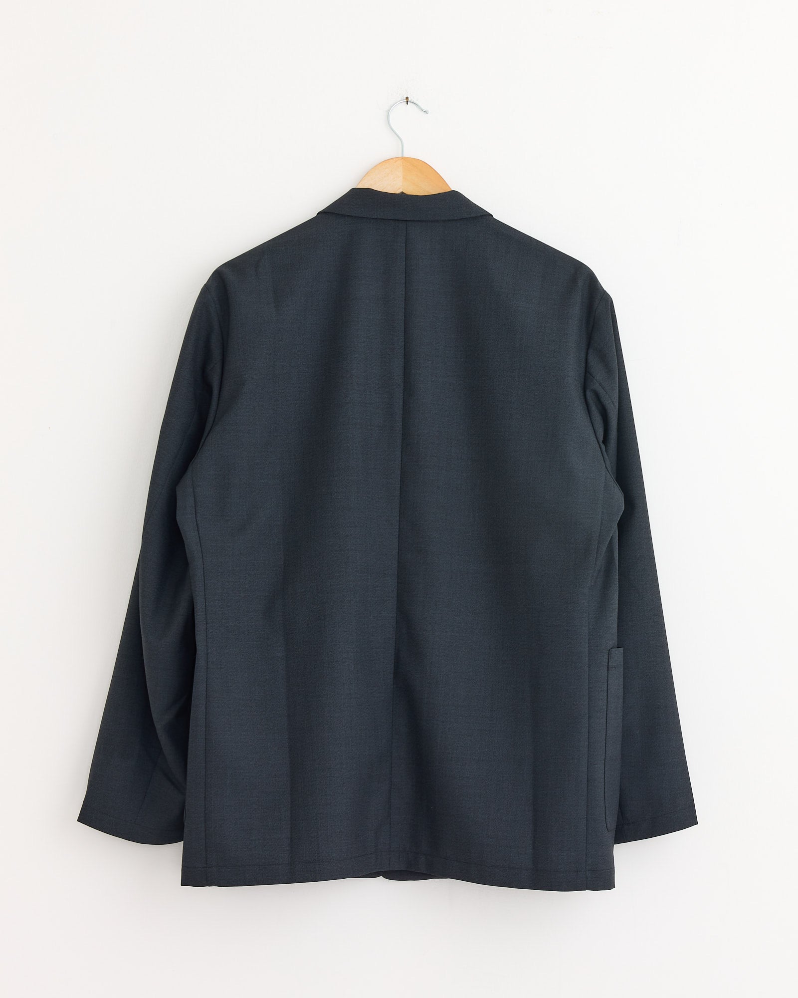 The Relaxed 2B Jacket in Charcoal by Still By Hand, featuring a tailored silhouette, is displayed on a wooden hanger against a plain white wall with the back view visible.