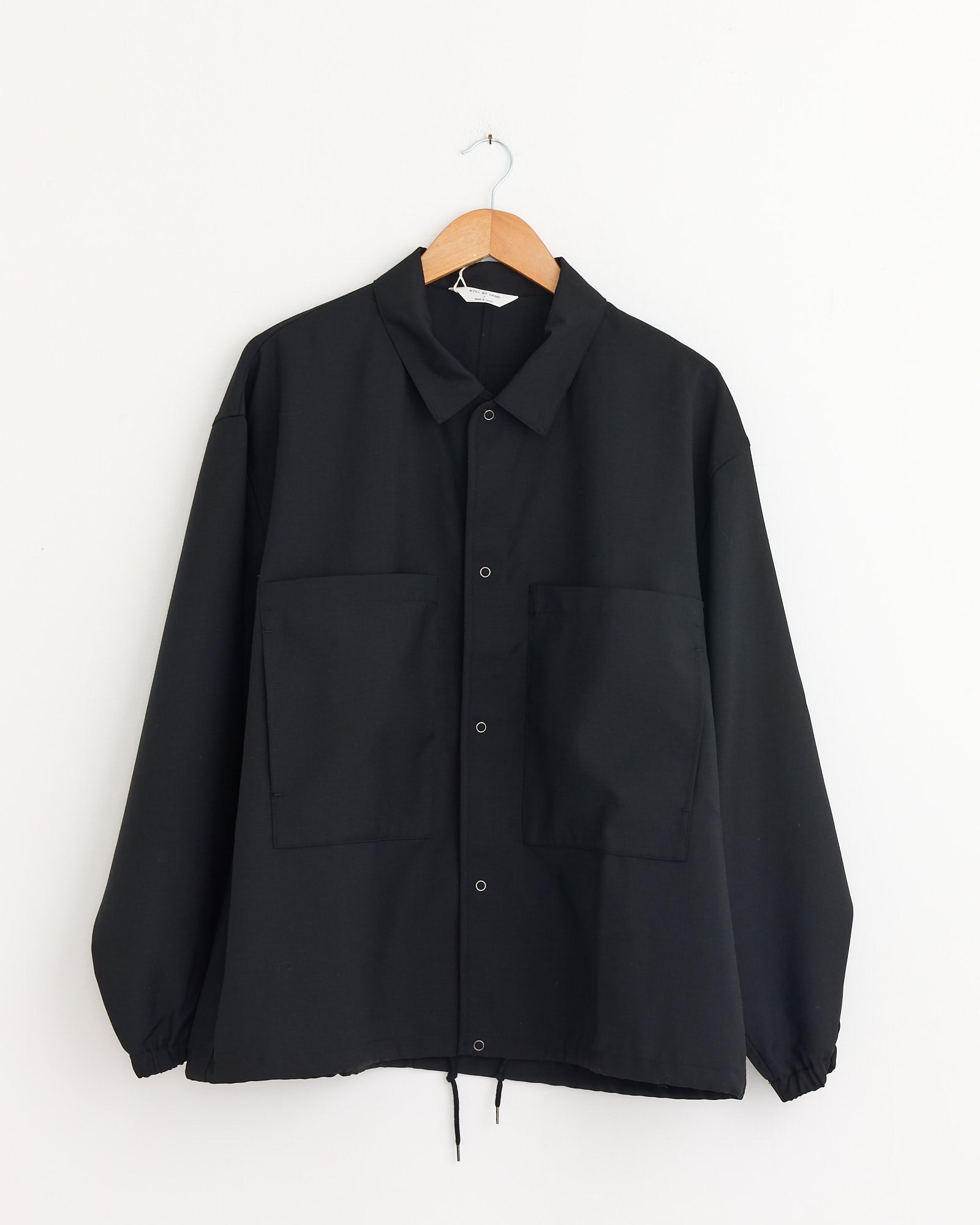The Still By Hand Double Pocket Shirt Jacket in Ink Black, made in Japan, features long sleeves, a button-up front, and two large front pockets. It is displayed on a wooden hanger against a plain white wall.