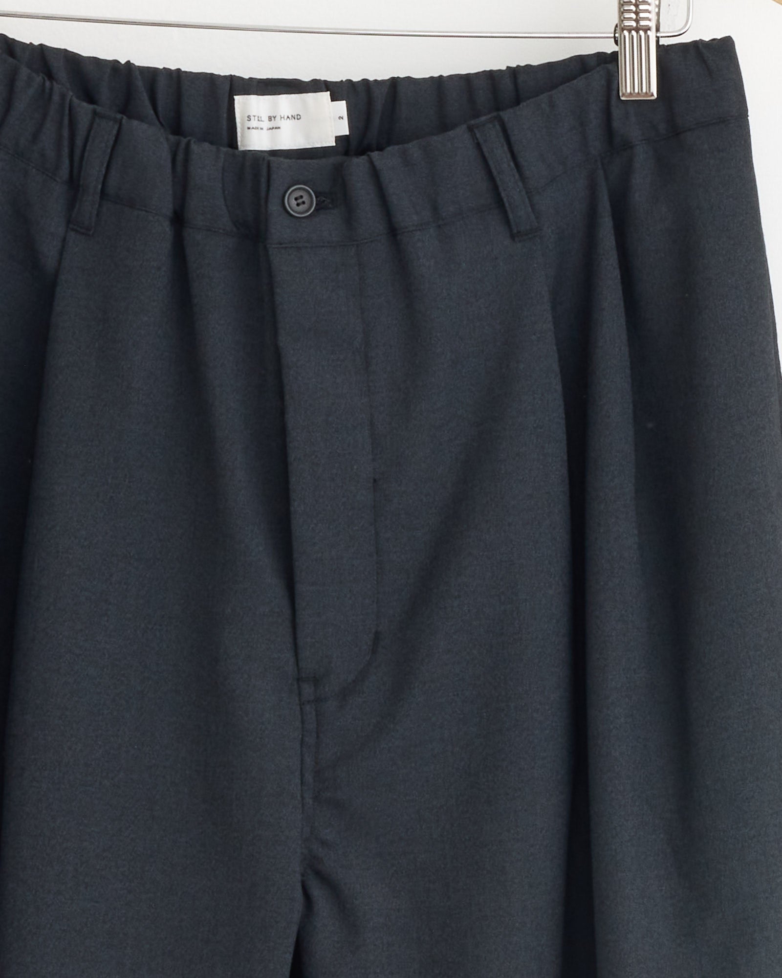 Close-up of the Relaxed 2 Tuck Pant in Charcoal by Still By Hand, made in Japan, hanging on a hanger and showing the waistband, button, zipper fly, and part of the inner label.