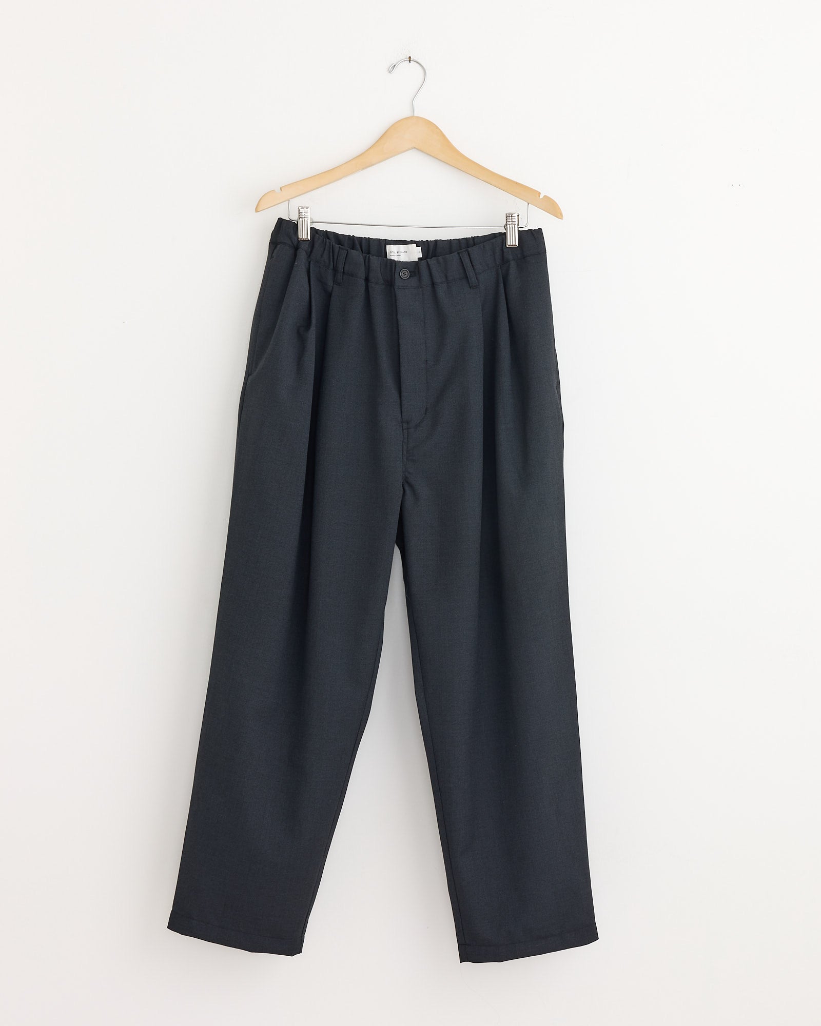 The Still By Hand Relaxed 2 Tuck Pant in Charcoal is a loose-fitting, wool-blend pant with an elastic waistband and button, shown on a wooden hanger against a plain white wall.