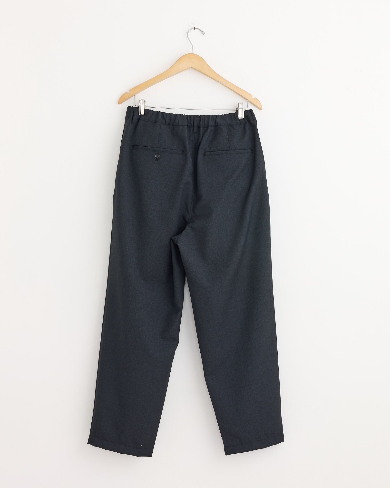 Still By Hand’s Relaxed 2 Tuck Pant in Charcoal features a wool blend, elastic waistband, and dual back pockets (one with a button). Made in Japan for quality and comfort. Shown hanging on a wooden hanger against a plain white wall.