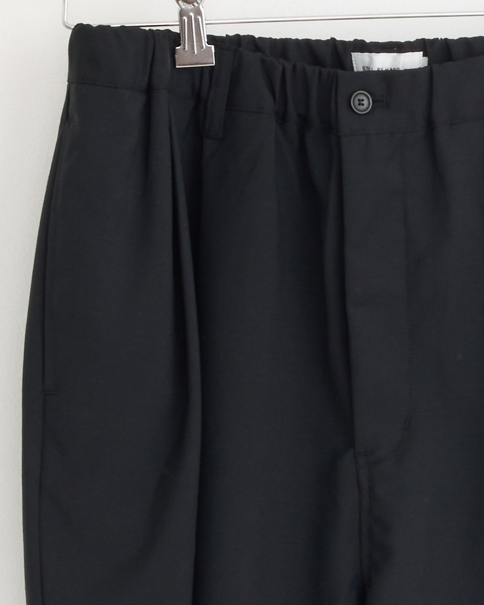 Close-up of the waistband and upper section of the Still By Hand Relaxed 2 Tuck Pant in Ink Black, featuring an elastic waistband, button, and pleated wool fabric, hanging on a silver clip hanger.