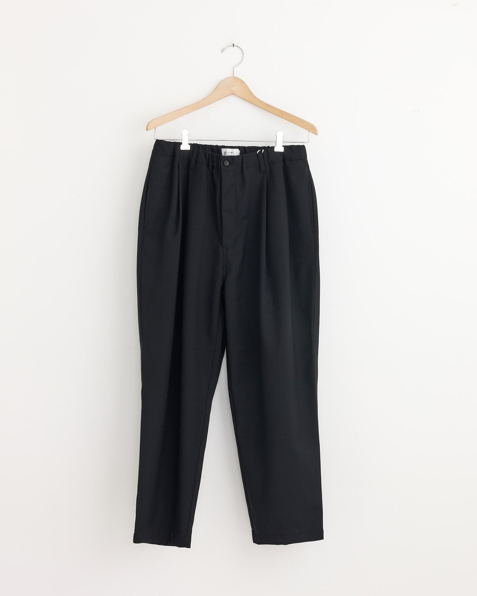 The Relaxed 2 Tuck Pant in Ink Black by Still By Hand features a tapered leg, elastic waistband, and pleats, shown hanging on a wooden hanger against a plain white wall.