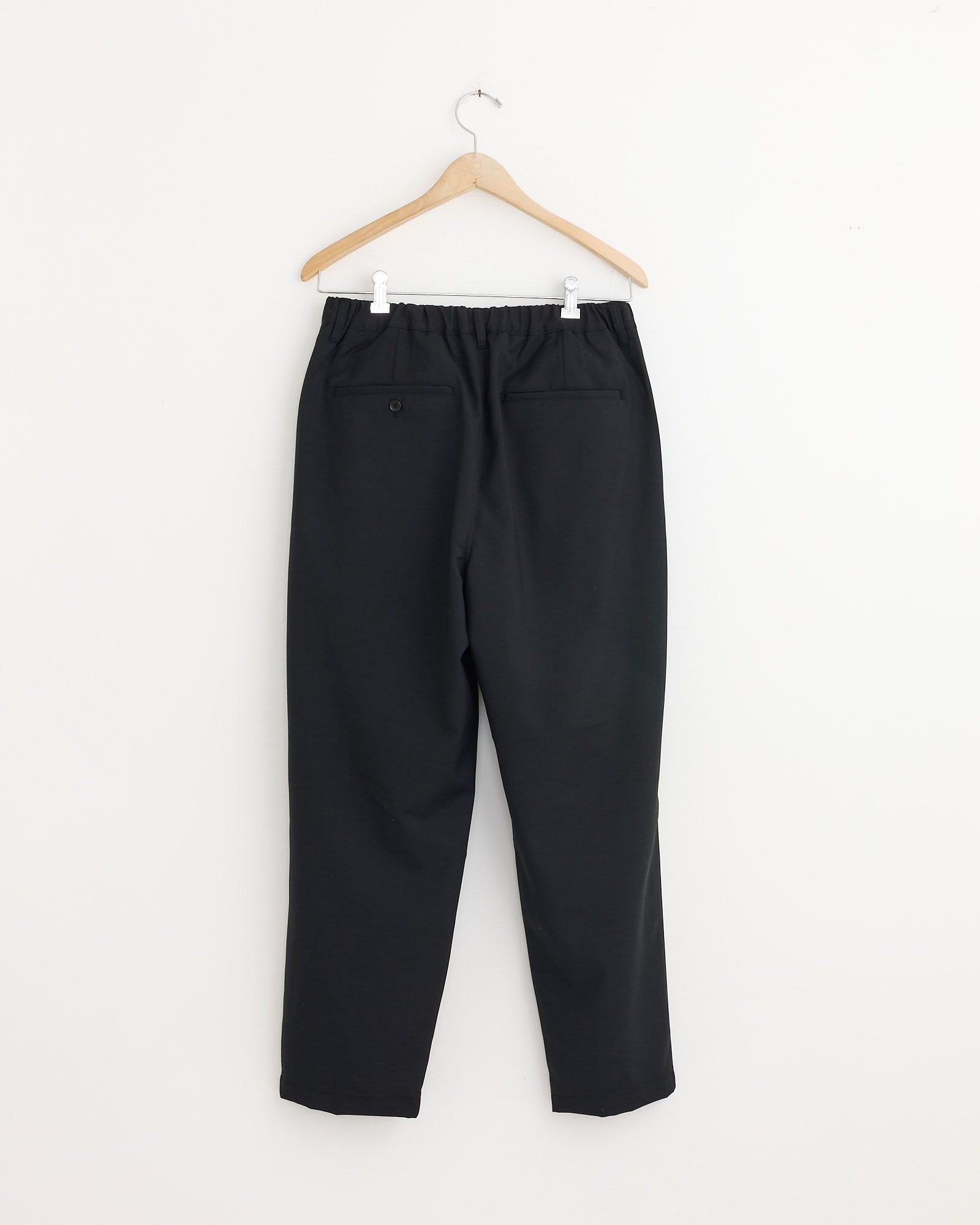 The Still By Hand Relaxed 2 Tuck Pant in Ink Black features an elastic waistband, two back pockets, and a tapered leg. Made in Japan, shown on a wooden hanger against a plain white wall.