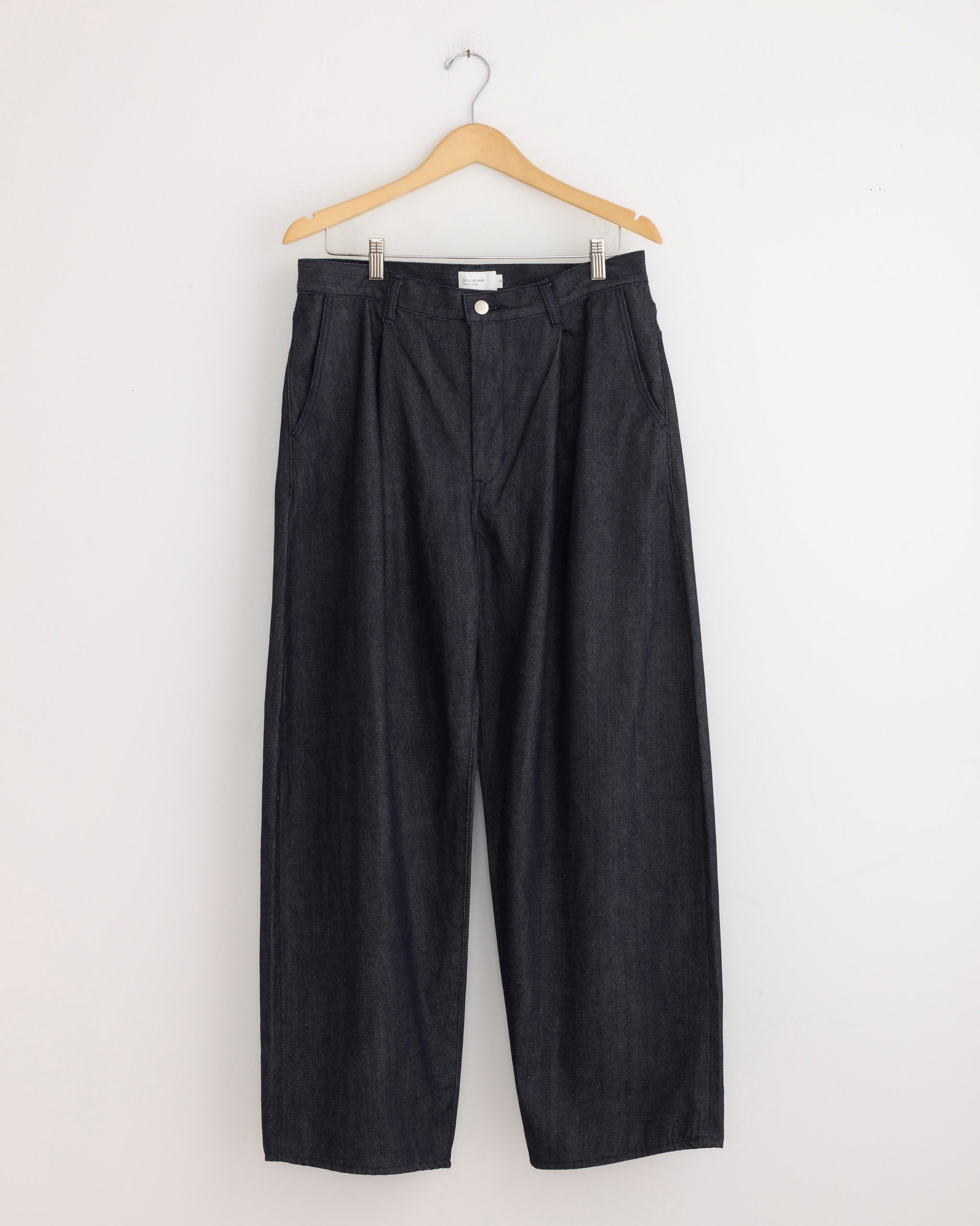 The Deep Tuck Wide Denim Pant in Navy by Still By Hand hangs on a wooden hanger against a white wall, featuring a wide-leg, high-waisted fit, front pockets, and button closure. Expertly crafted in Japan.