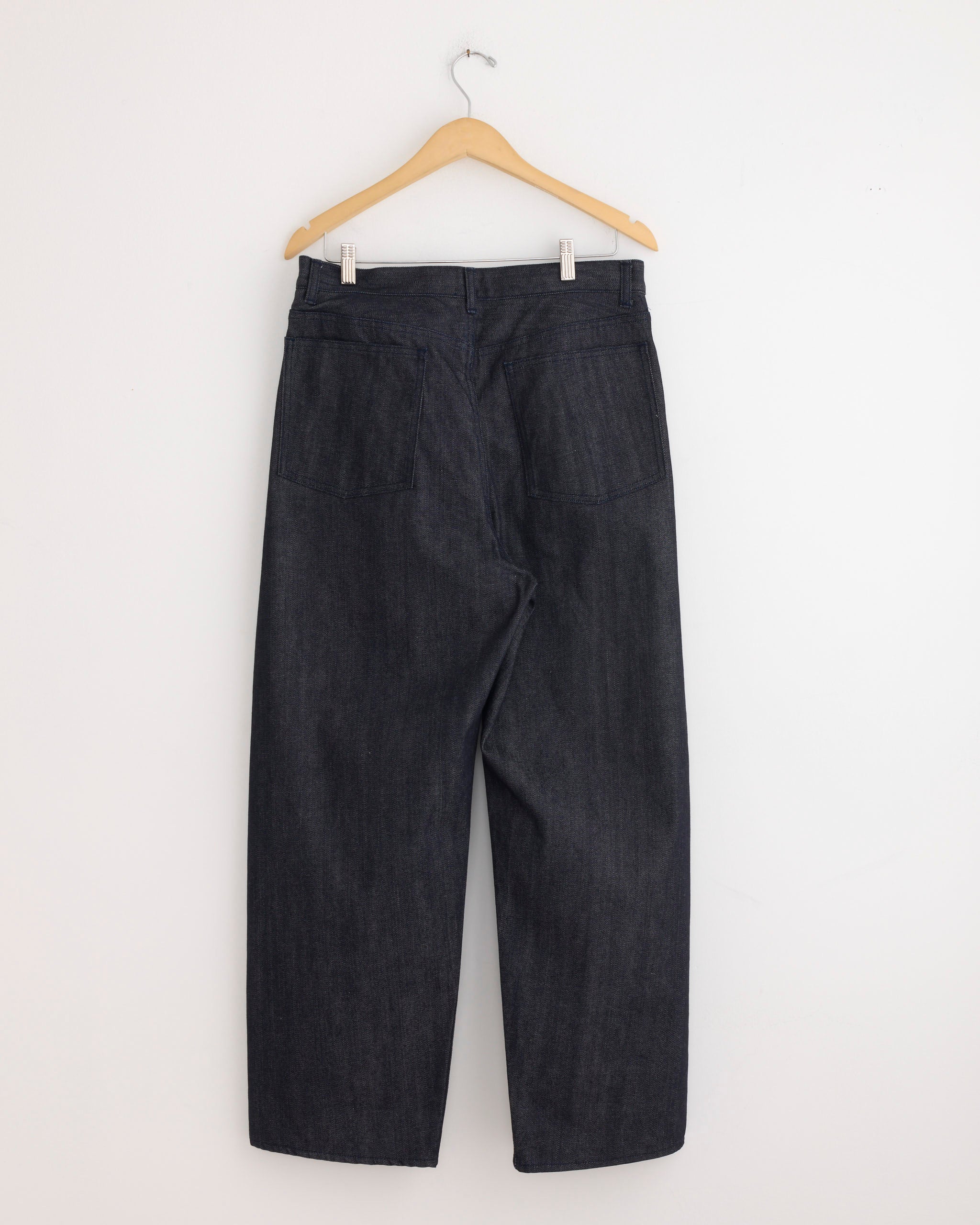 Still By Hand's Deep Tuck Wide Denim Pant in Navy, crafted from premium Kojima Okayama denim, is displayed on a wooden hanger against a white wall, showing the back with two large rear pockets and a simple waistband.