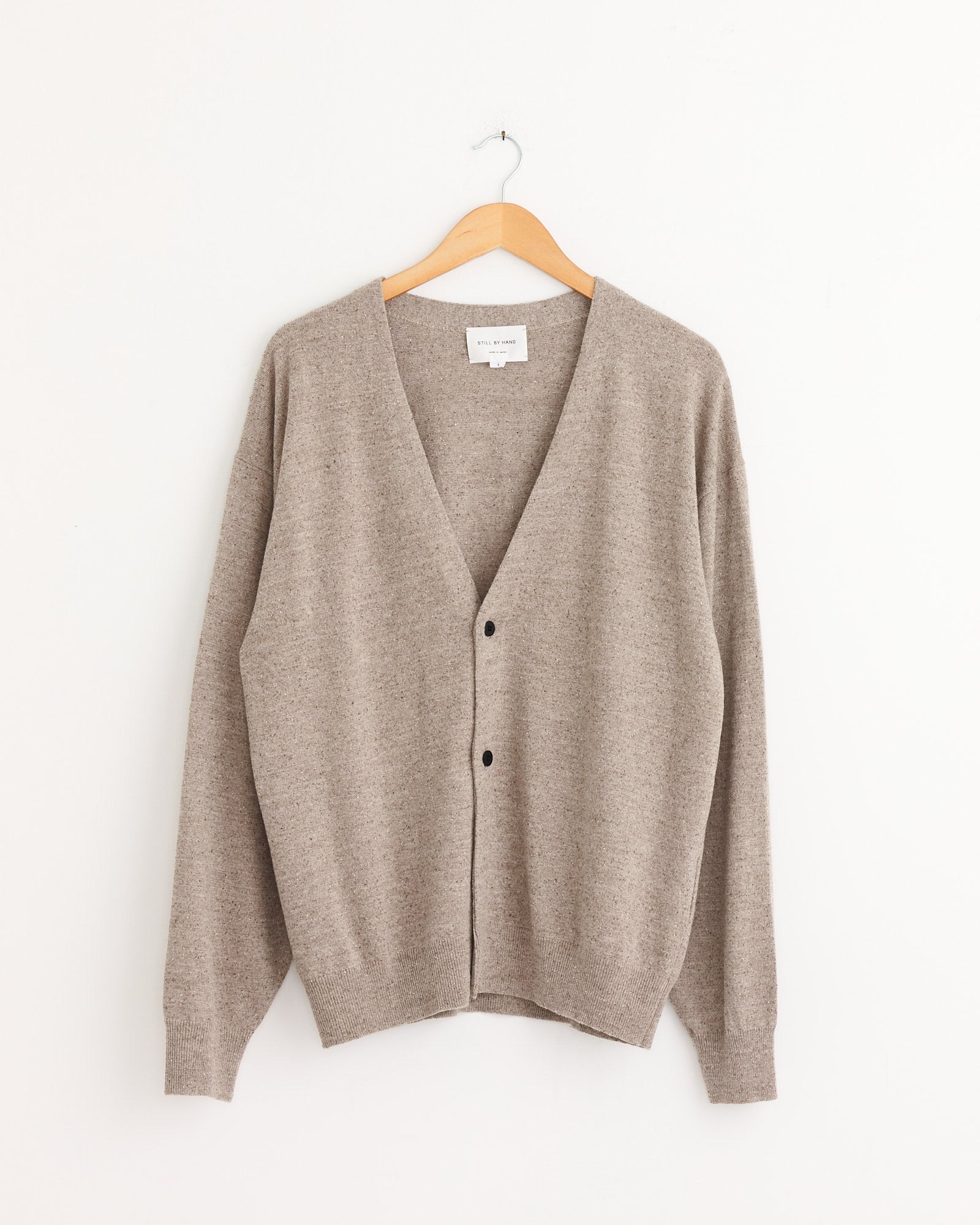 The Still By Hand Silk Mixed 2B Cardigan in Sand Beige, a soft jersey long-sleeve V-neck with two buttons, hangs on a wooden hanger against a plain white wall.