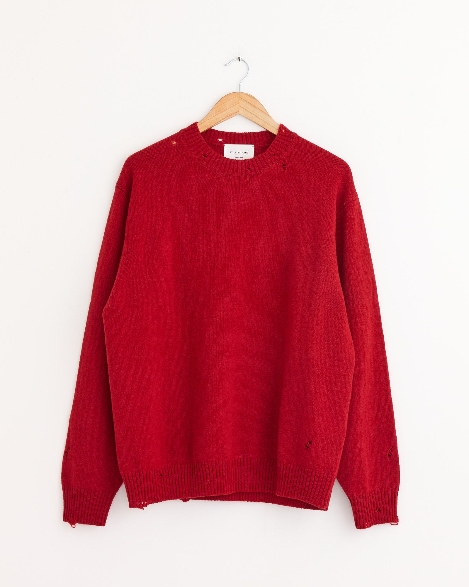 The Punched Shetland Sweater in Vermilion Red by Still By Hand hangs on a wooden hanger, showcasing its crew neck and ribbed details. Made in Japan, this classic wool sweater offers timeless style and comfort against a plain white background.