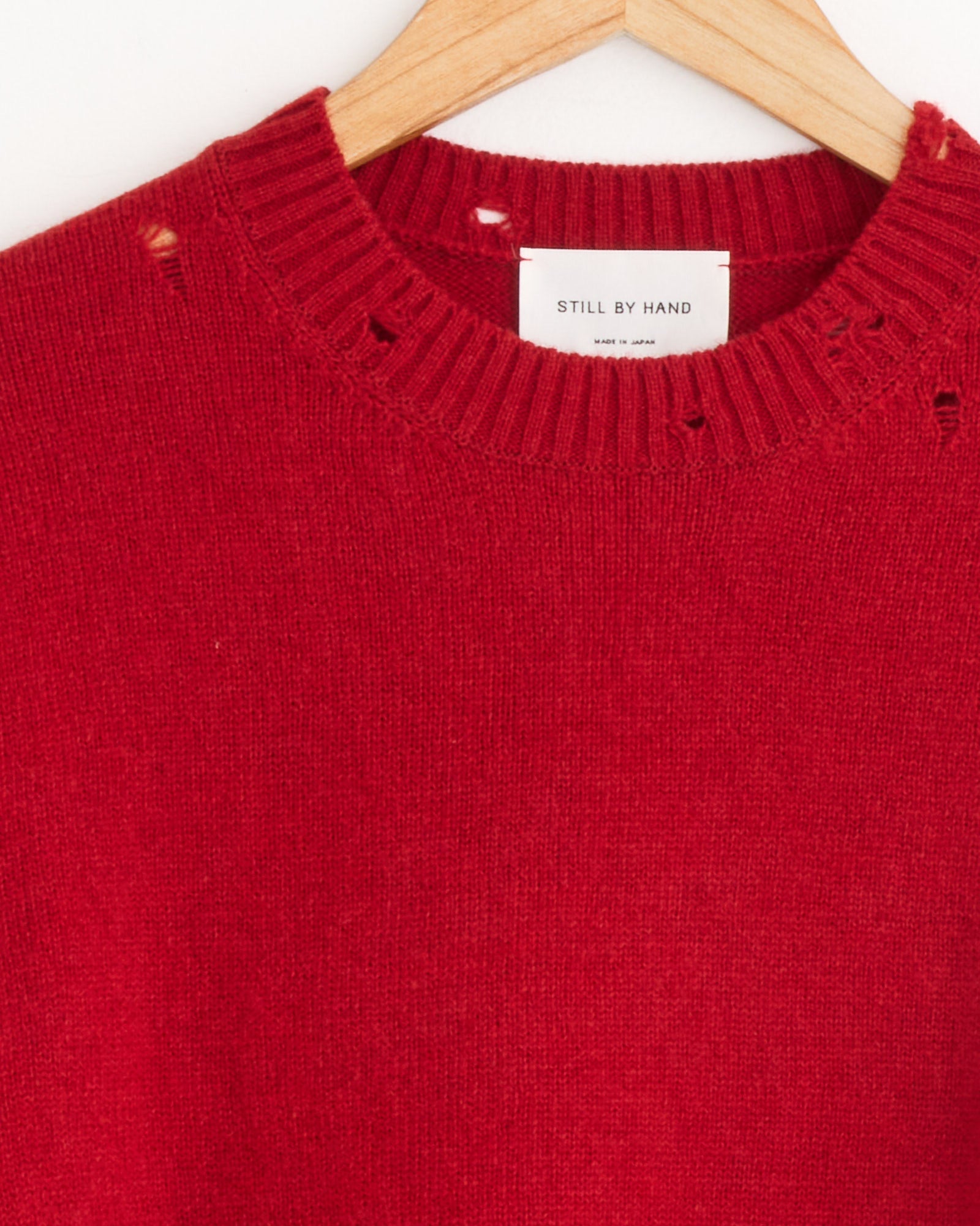 A close-up of the Still By Hand Punched Shetland Sweater in Vermilion Red on a wooden hanger shows minor holes near the collar and a white Still By Hand label, highlighting the brand’s made-in-Japan craftsmanship.