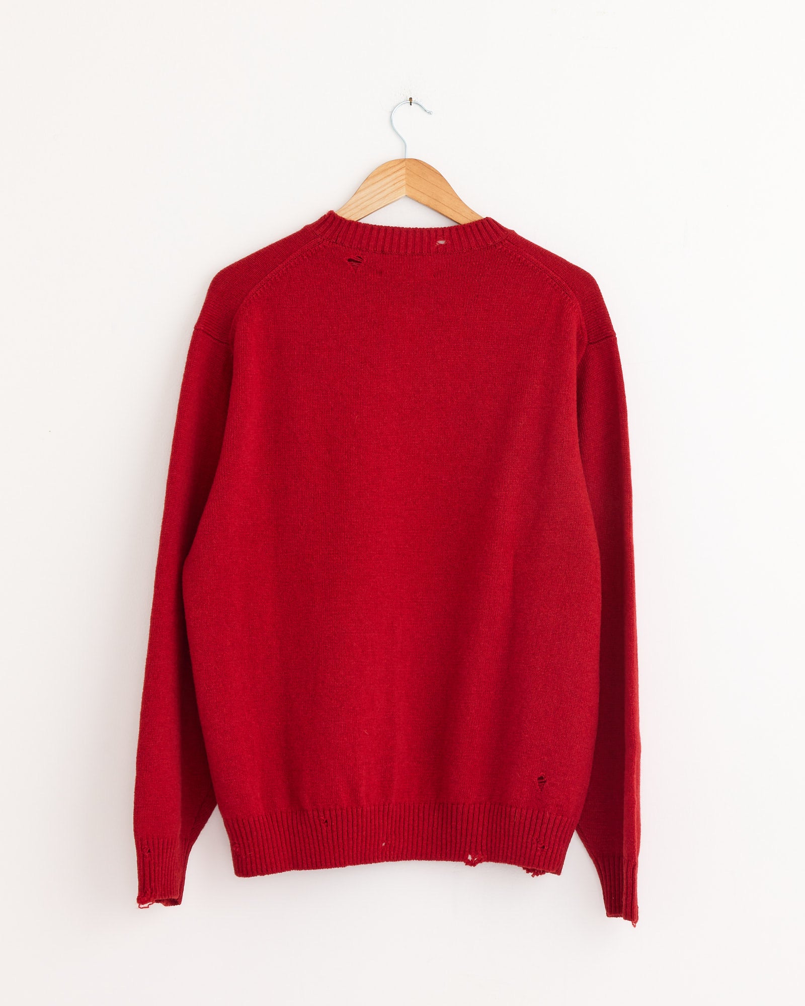 The Punched Shetland Sweater in Vermilion Red by Still By Hand hangs on a wooden hanger against a white wall. Made in Japan, it has a ribbed collar, cuffs, and hem, with slight wear and small holes near the collar and hem.