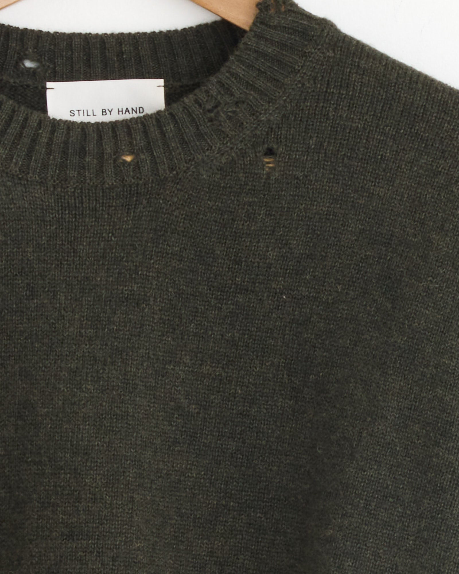 A close-up of the Still By Hand Punched Shetland Sweater in Khaki Green on a hanger shows small holes by the ribbed neckline and a label inside, marking this Japanese-made piece.
