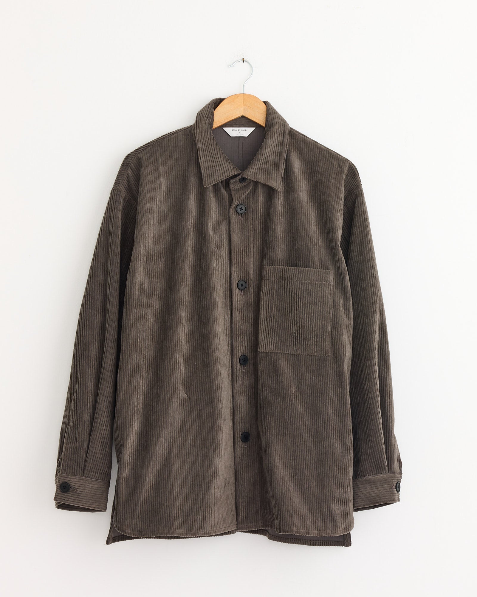 The Still By Hand Corduroy Shirt Jacket in Khaki Brown, crafted in Japan from 100% cotton, features long sleeves and a single chest pocket, and is displayed on a wooden hanger against a plain white wall.