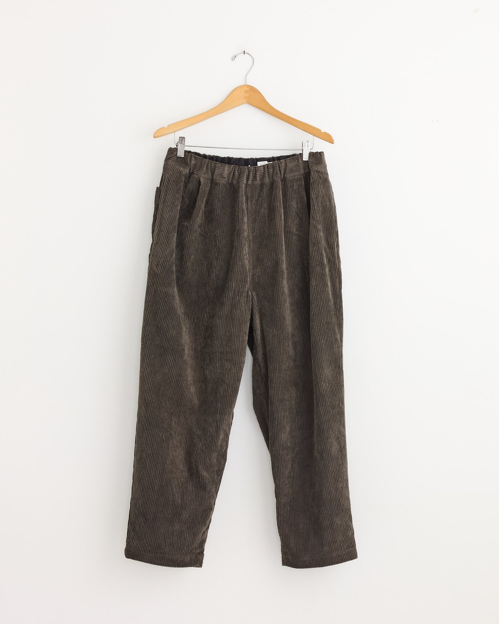 The Still By Hand Corduroy Easy Pant in Khaki Brown, crafted in Japan, features an elastic waistband and is displayed on a wooden hanger against a plain white wall.