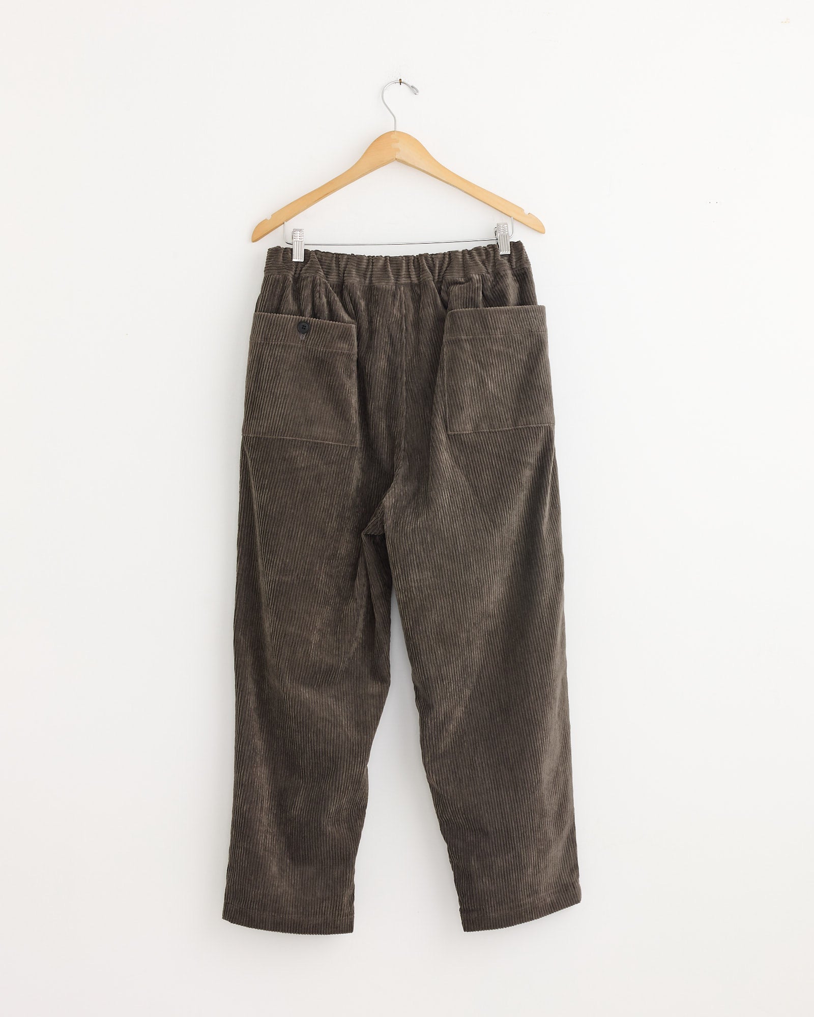The Still By Hand Corduroy Easy Pant in Khaki Brown features an elastic waistband and two large front pockets. Shown hanging on a wooden hanger against a plain white wall. Made in Japan.