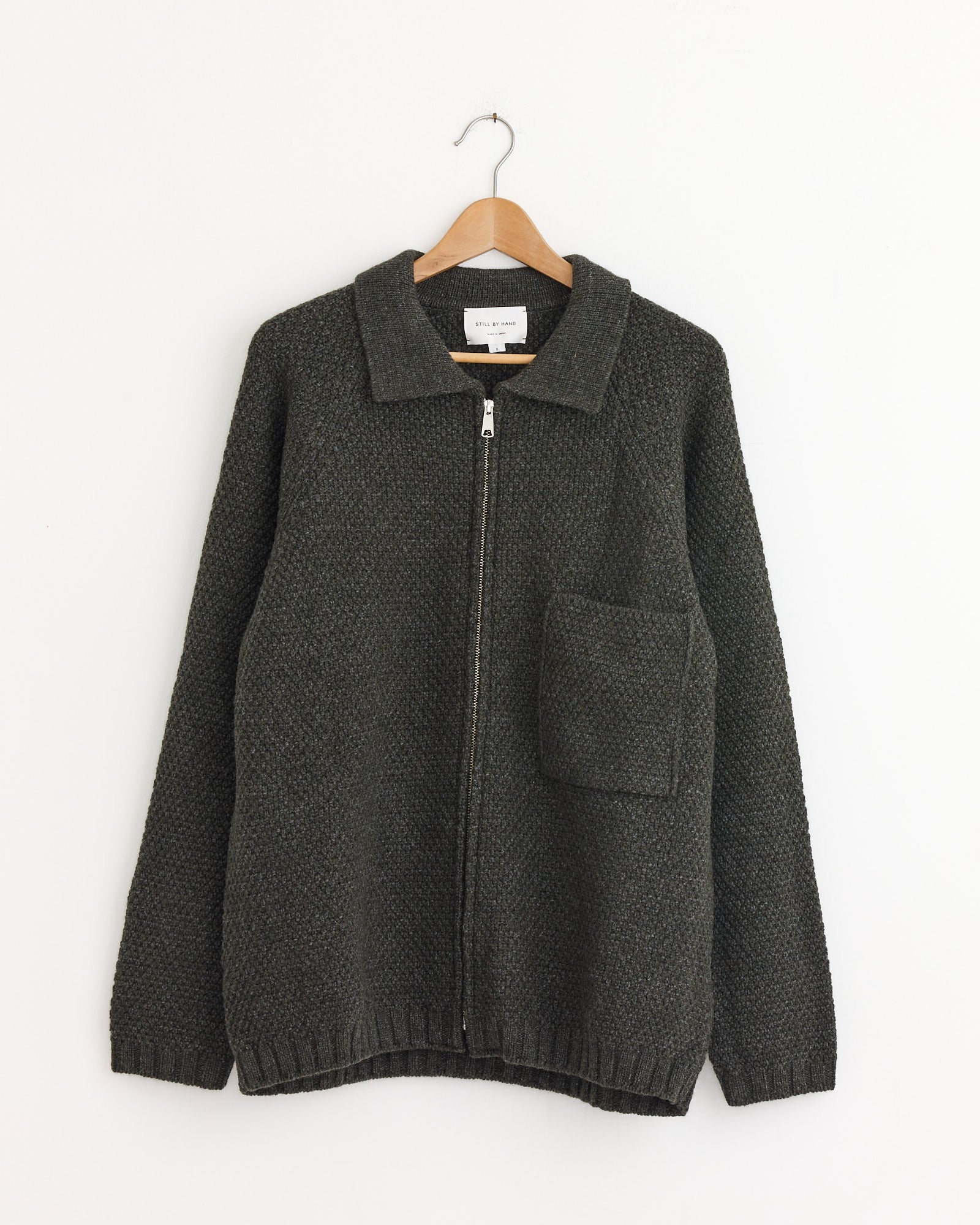 The 5G Knitted Blouson in Melange Khaki by Still By Hand hangs on a wooden hanger with a large front pocket, displayed against a plain white wall.