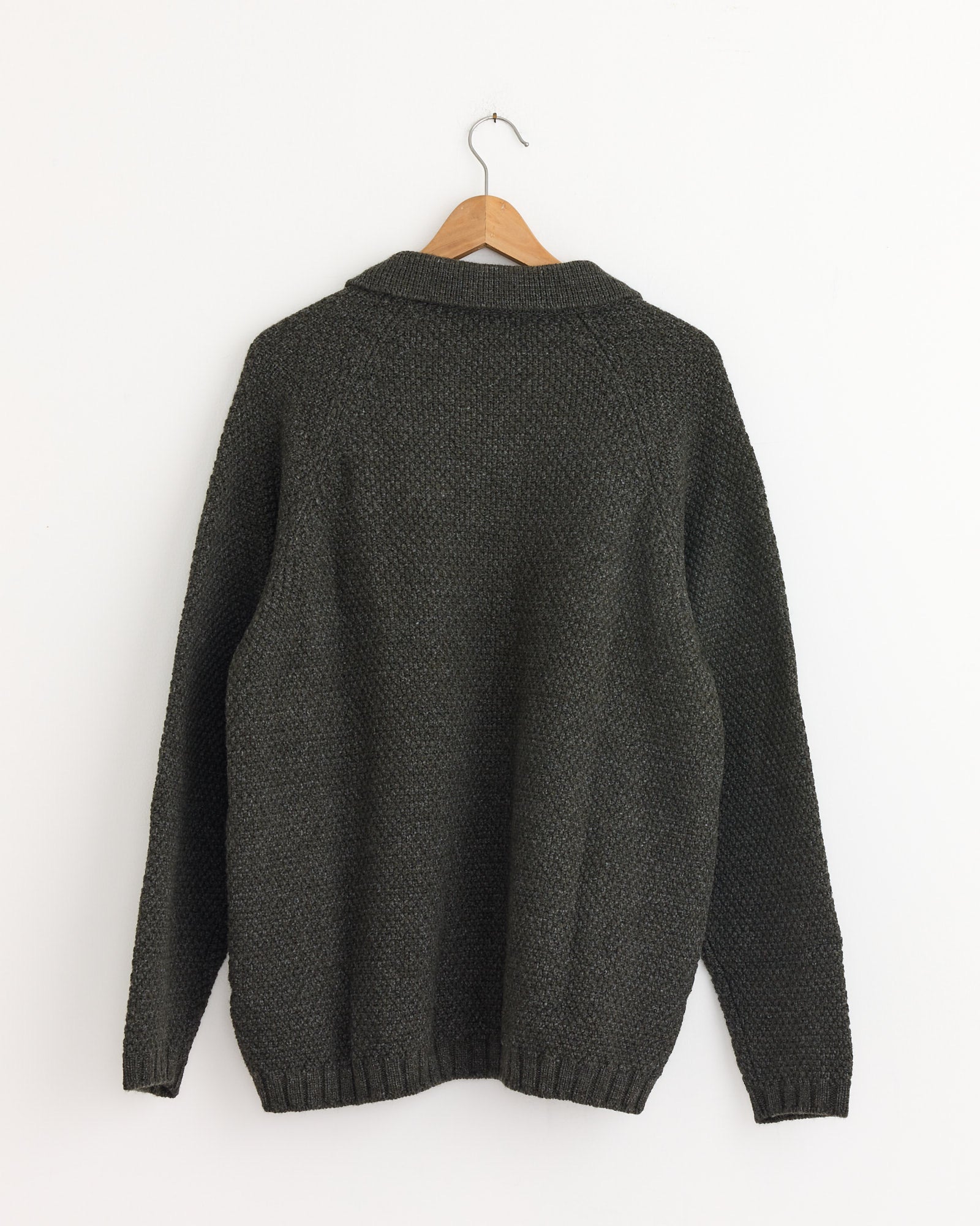 The Still By Hand 5G Knitted Blouson in Melange Khaki hangs on a wooden hanger against a plain white wall, shown from the back. Made in Japan.