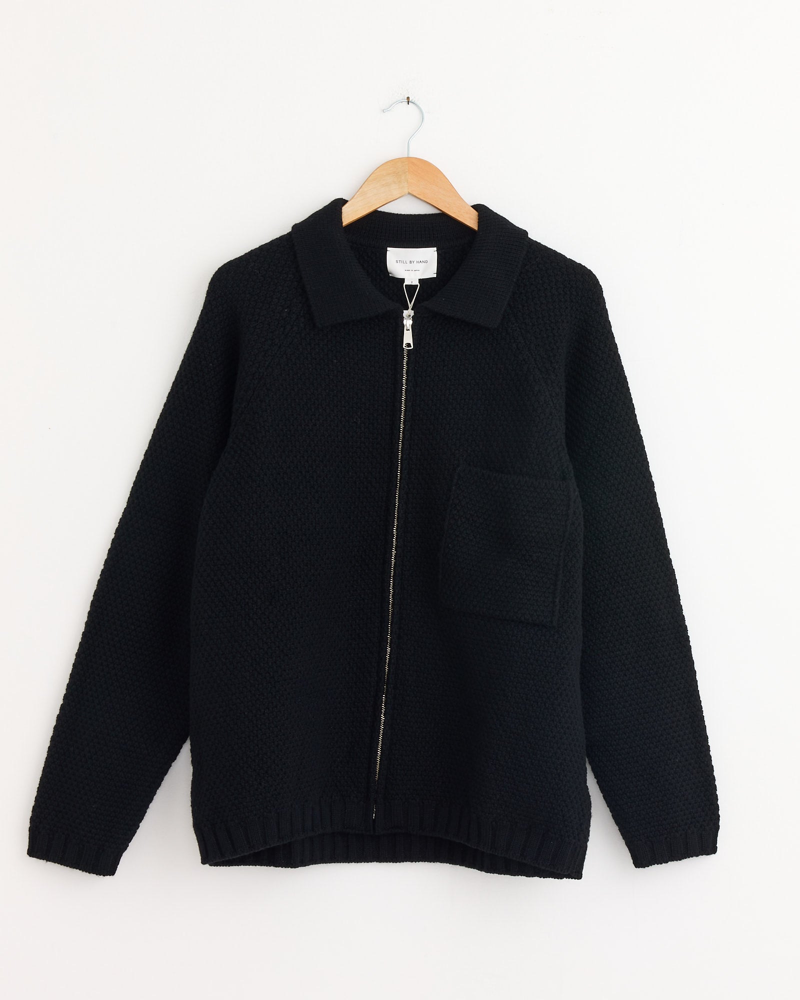 The Still By Hand 5G Knitted Blouson in Black, featuring a collar and single front pocket, is displayed on a wooden hanger against a white background.