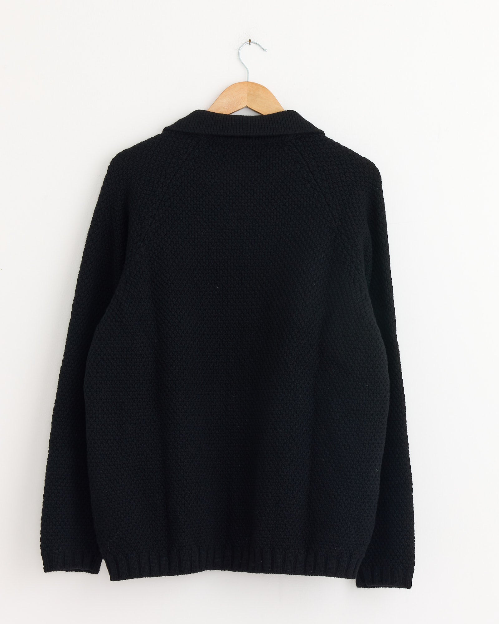 The Still By Hand 5G Knitted Blouson in Black is displayed from the back on a wooden hanger against a white wall.