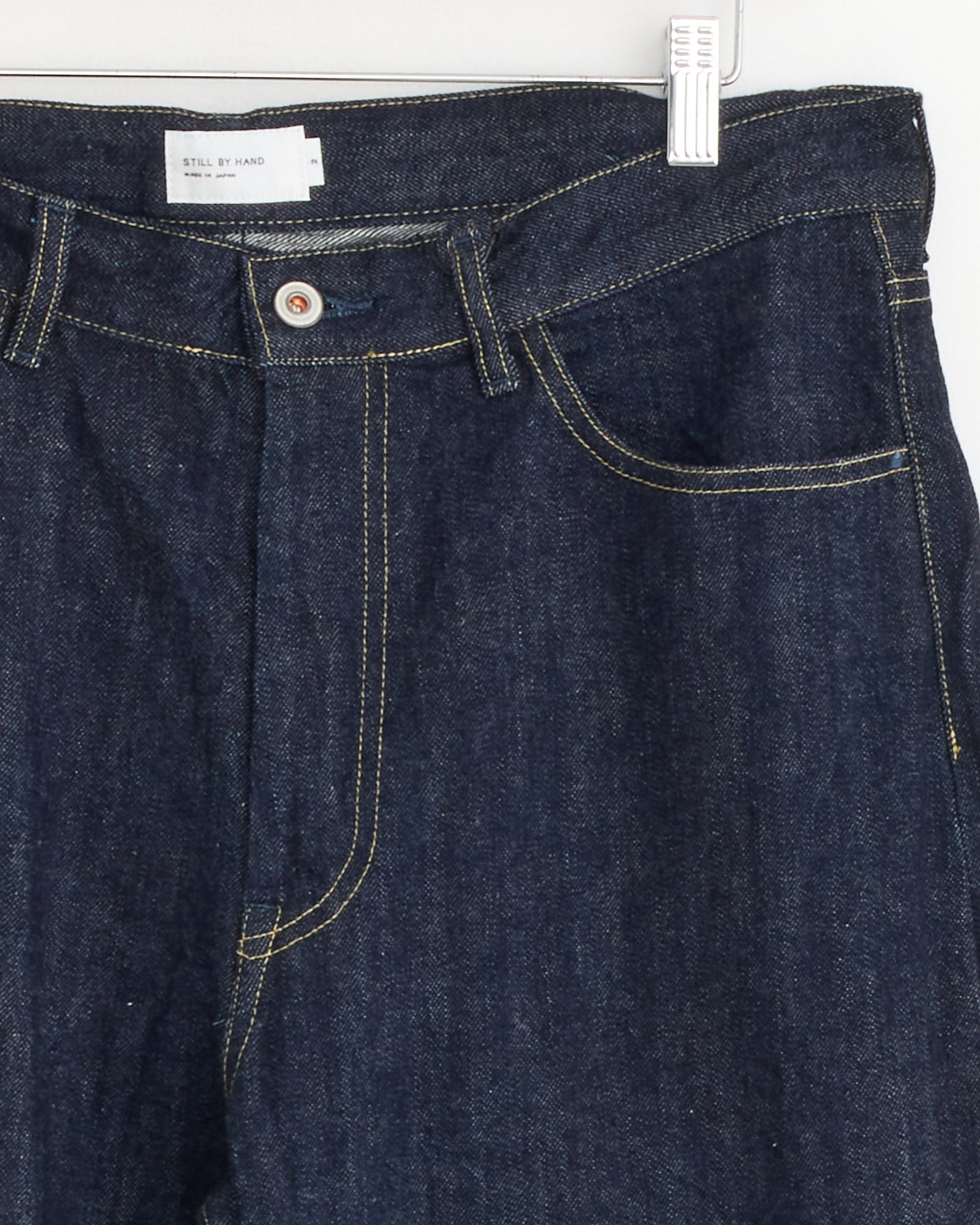 Close-up of Still By Hand's 5 Pocket Denim Pant in Navy, featuring white selvedge denim, yellow stitching, a silver button, and a small white waistband label, hanging on a clip against a white background.