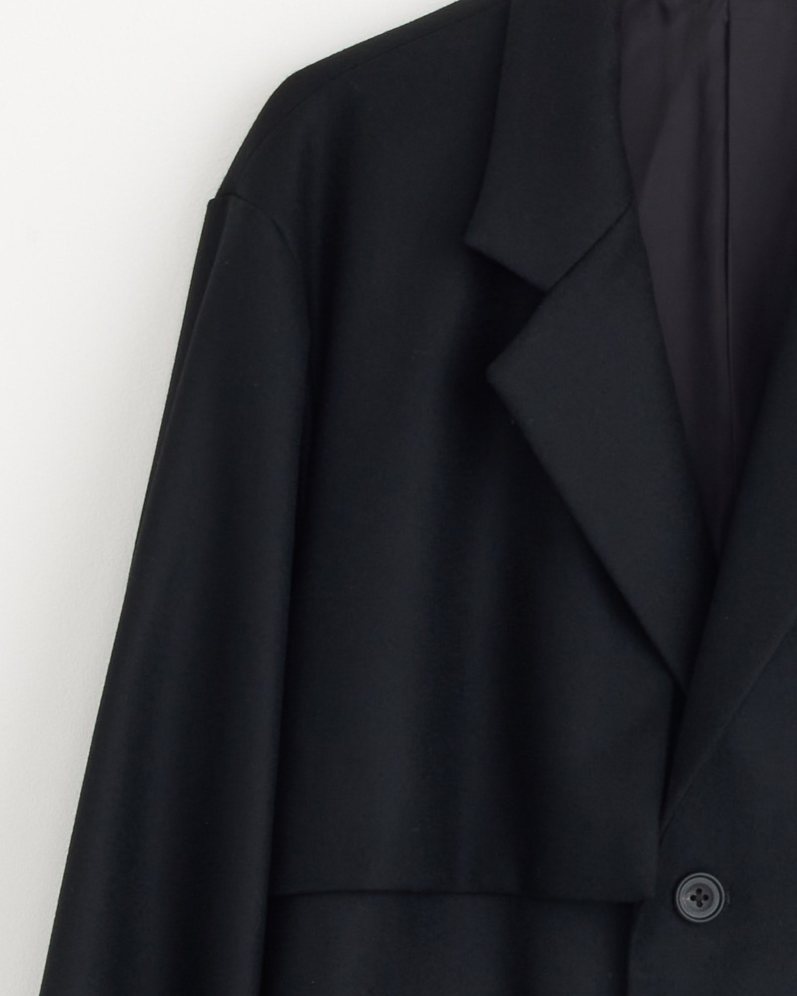 A close-up of the ssstein 1211 Coat in Black, a minimalist wool melton Chester coat with a notched lapel and single visible button, hanging against a plain white background.