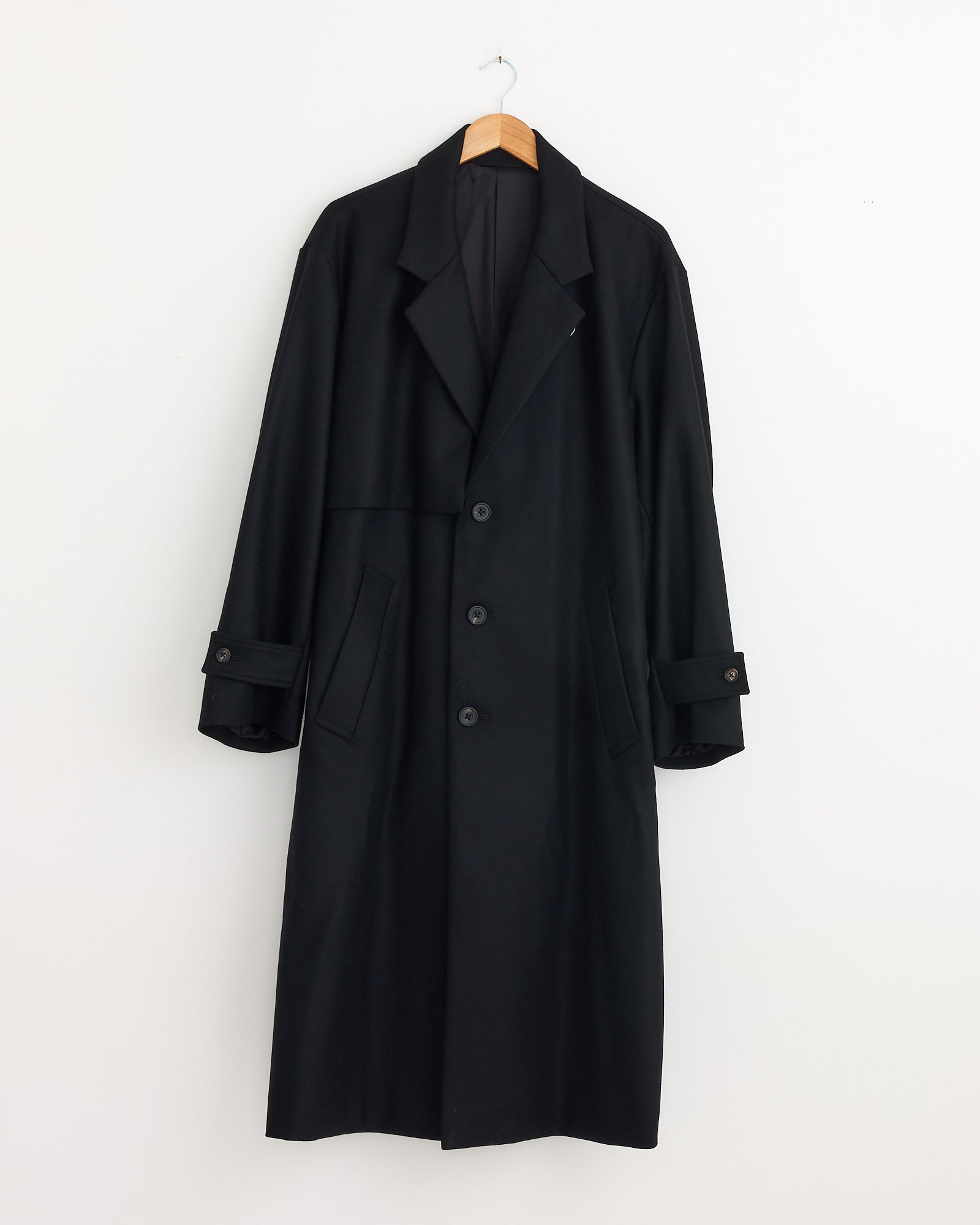 The ssstein 1211 Coat in Black—a minimalist wool melton coat with three buttons, notched lapels, and buttoned cuffs—is displayed on a wooden hanger against a plain white wall.