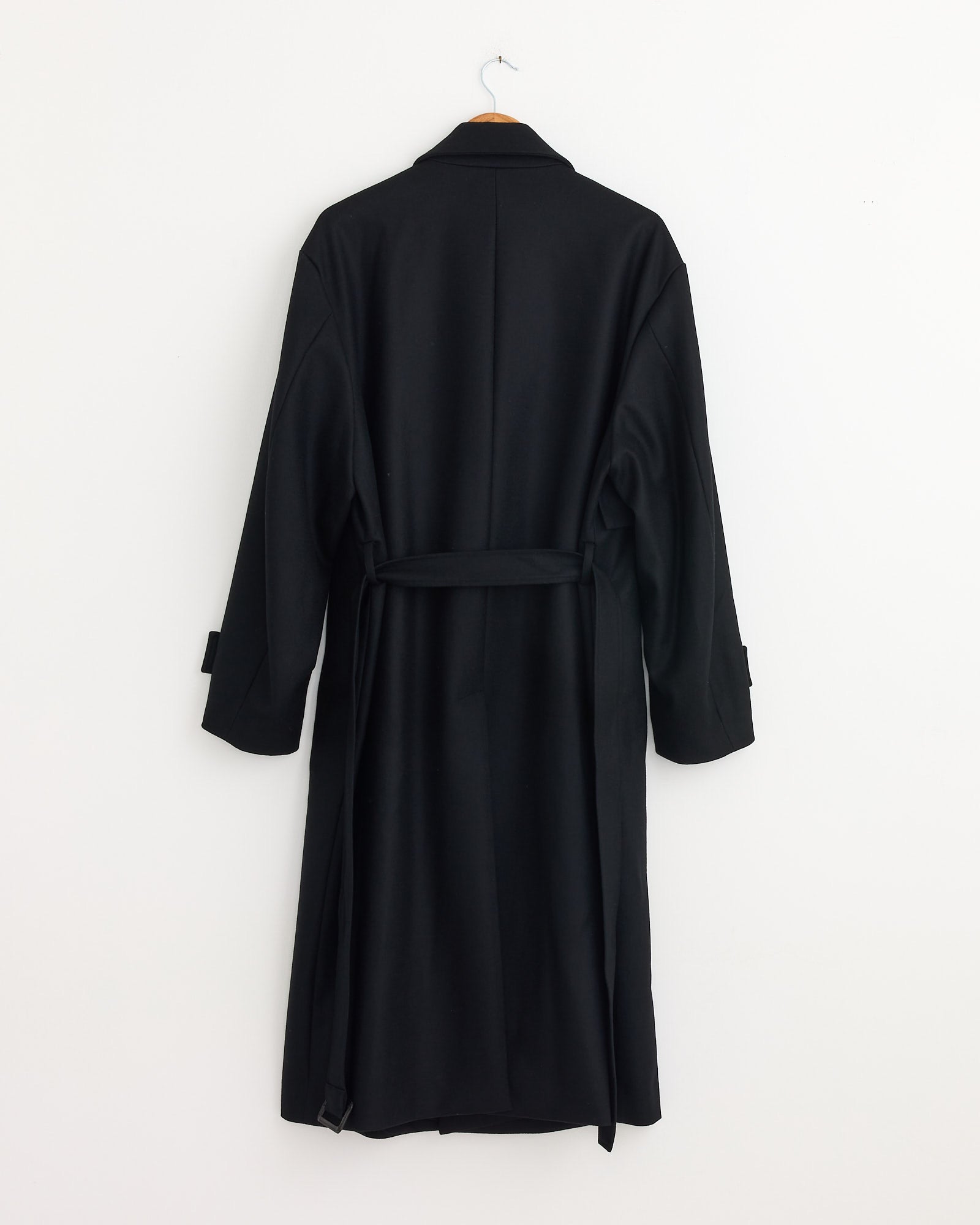 The ssstein 1211 Coat in Black, made from wool melton, hangs on a wooden hanger against a white wall. It features folded cuffs and a belt tied at the back.