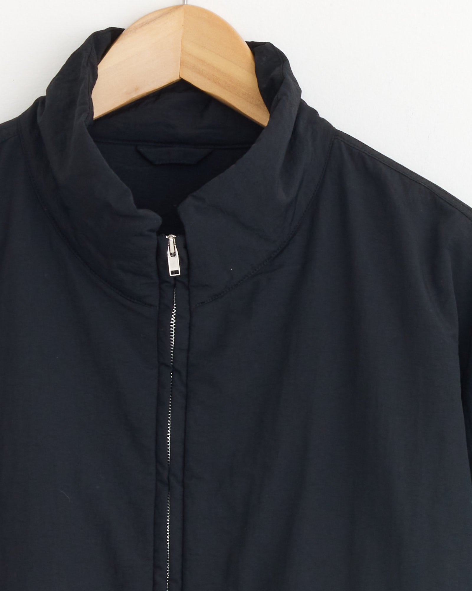 The 1215 Jacket in Black by ssstein, made from recycled nylon by OLMETEX and featuring a stand collar, hangs on a wooden hanger against a plain white background.