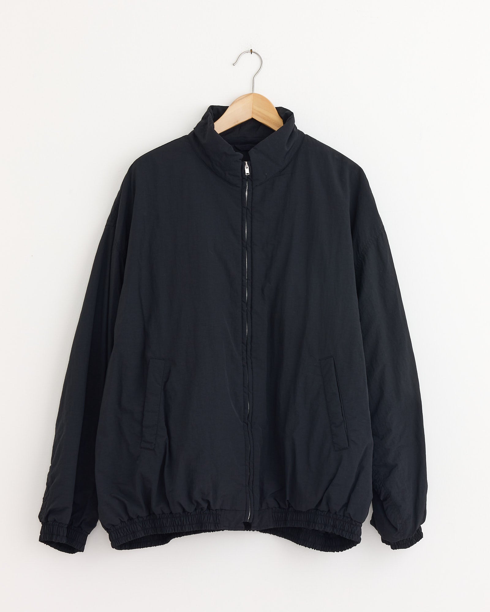 The 1215 Jacket in Black by ssstein is a padded, recycled nylon jacket with a high collar and elastic cuffs and hem, crafted by OLMETEX. Shown hanging on a wooden hanger against a white wall.