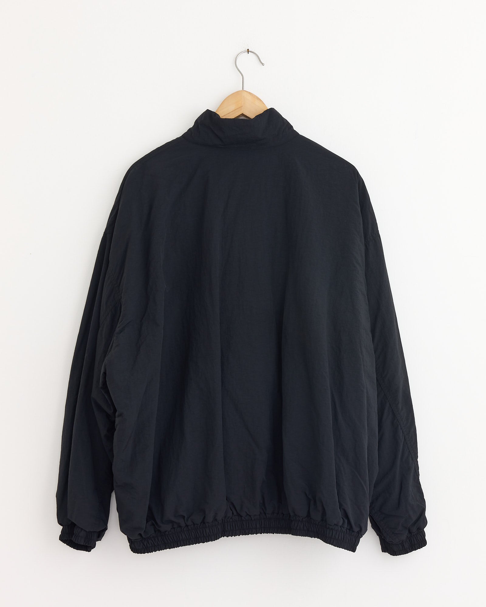 The ssstein 1215 Jacket in Black, made from recycled nylon with long sleeves and elastic cuffs and hem, is displayed on a wooden hanger against a white wall, shown from the back.