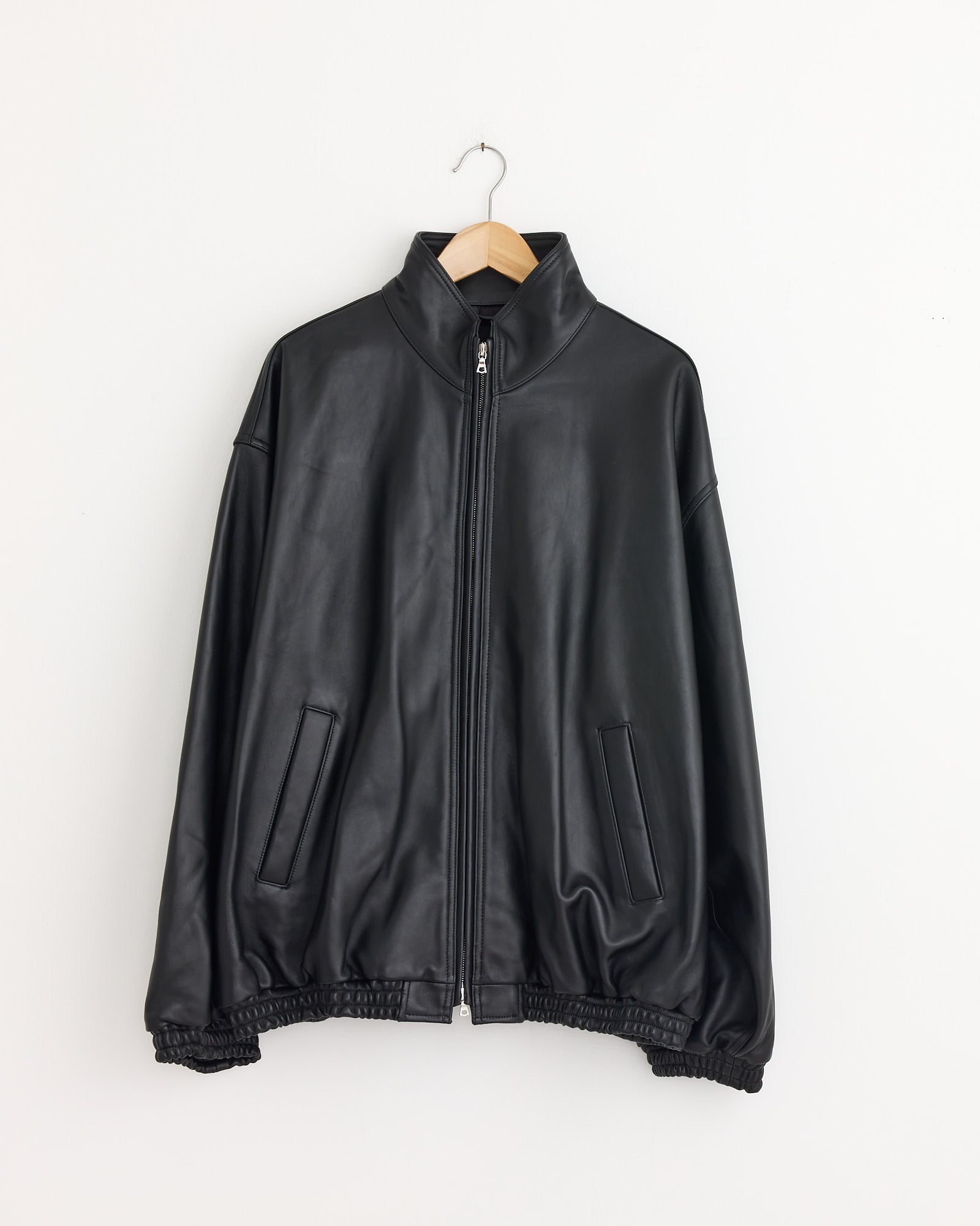 The ssstein 1220 Leather Jacket in Black, featuring a front zipper, stand collar, and two slanted side pockets, hangs on a wooden hanger against a white wall, showcasing timeless leather jacket style.