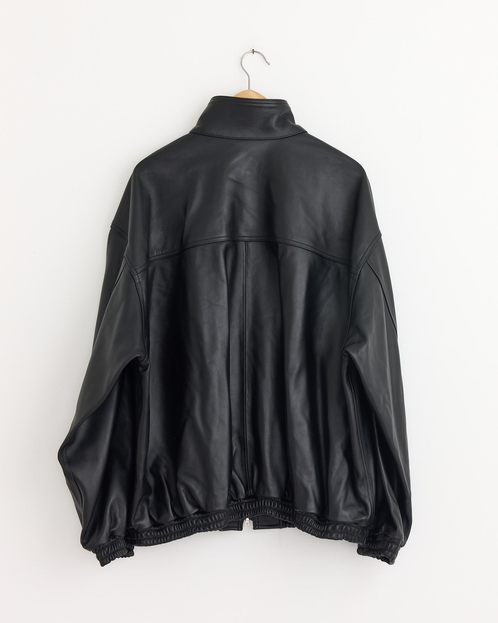 The ssstein 1220 Leather Jacket in Black, featuring a high collar and elastic cuffs, hangs on a wooden hanger against a plain white wall, displayed from the back.