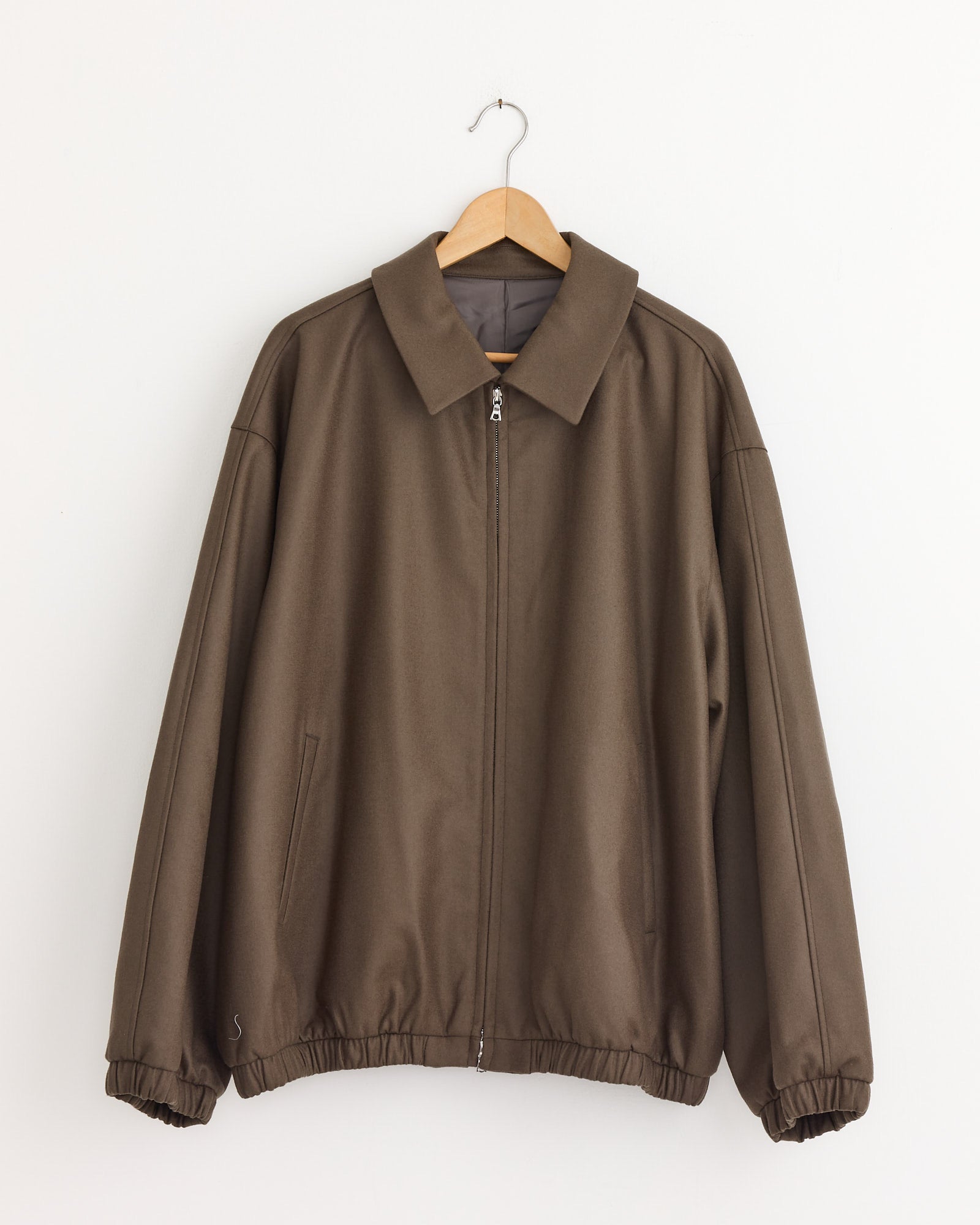 The ssstein Melton 1226 Jacket in Dark Beige, made from premium Super 120’s wool with a collar, elastic cuffs and hem, hangs on a wooden hanger against a plain white wall.