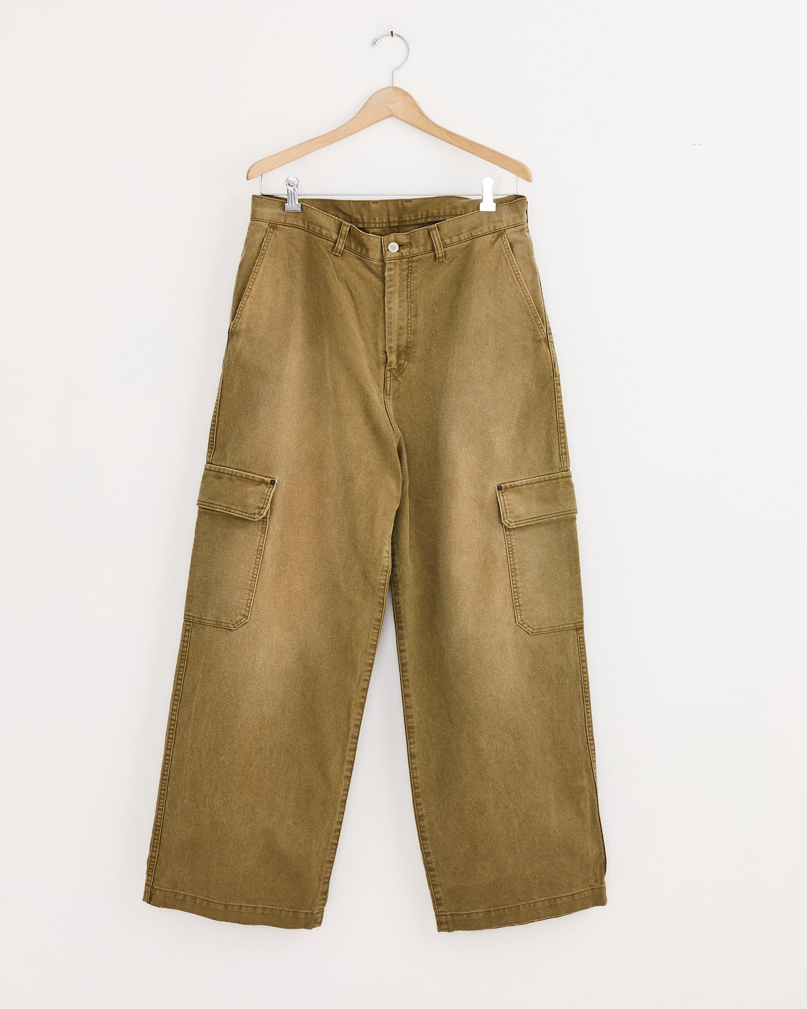 The ssstein Sulfur Dyed Trousers in Khaki are high-waisted, wide-leg cargo pants made from durable whipcord with large side pockets, shown on a wooden hanger against a plain white wall.