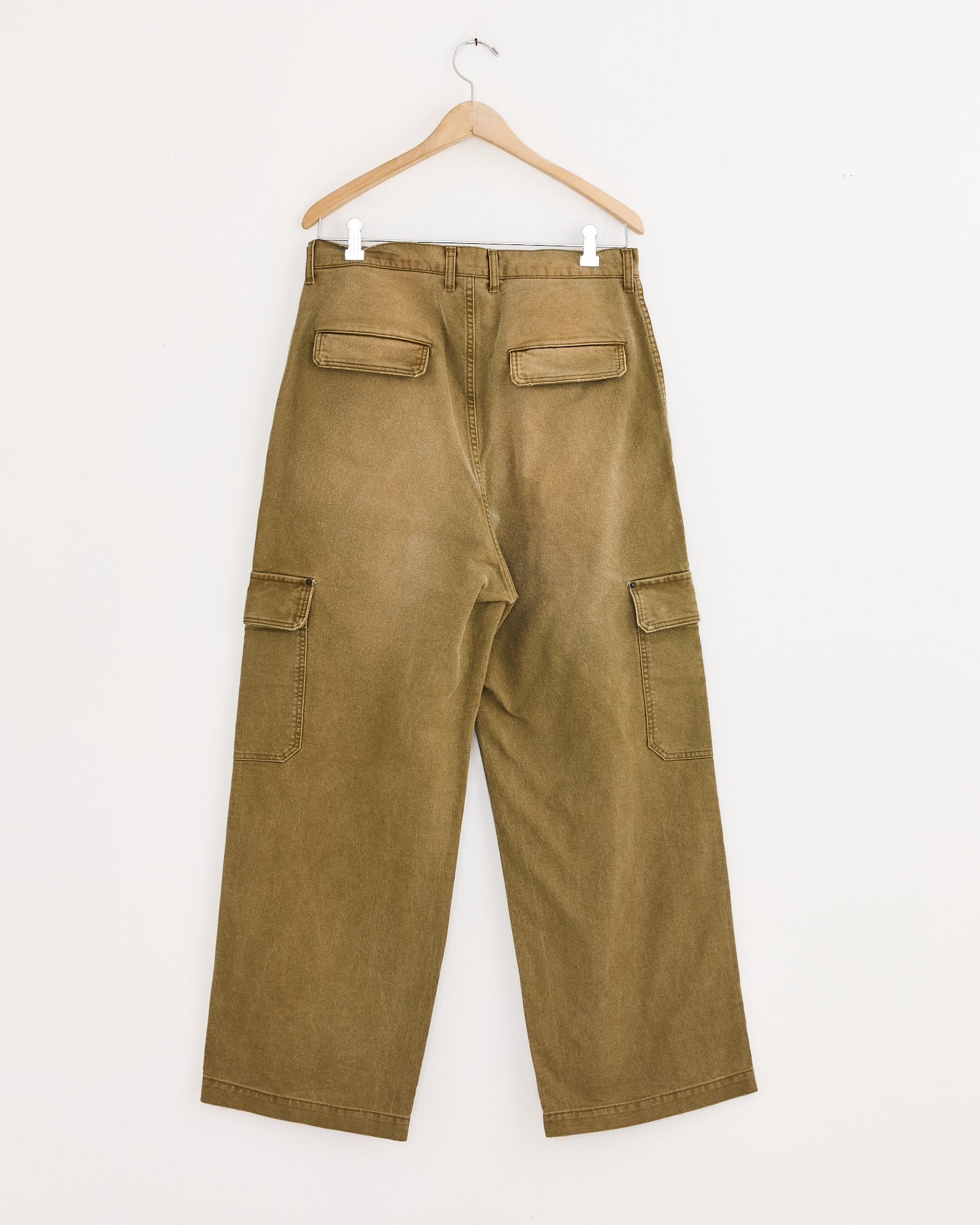 The ssstein Sulfur Dyed Trousers in Khaki hang on a wooden hanger against a white wall, displaying the back view with two flap pockets and two side cargo pockets, all crafted from durable whipcord fabric.