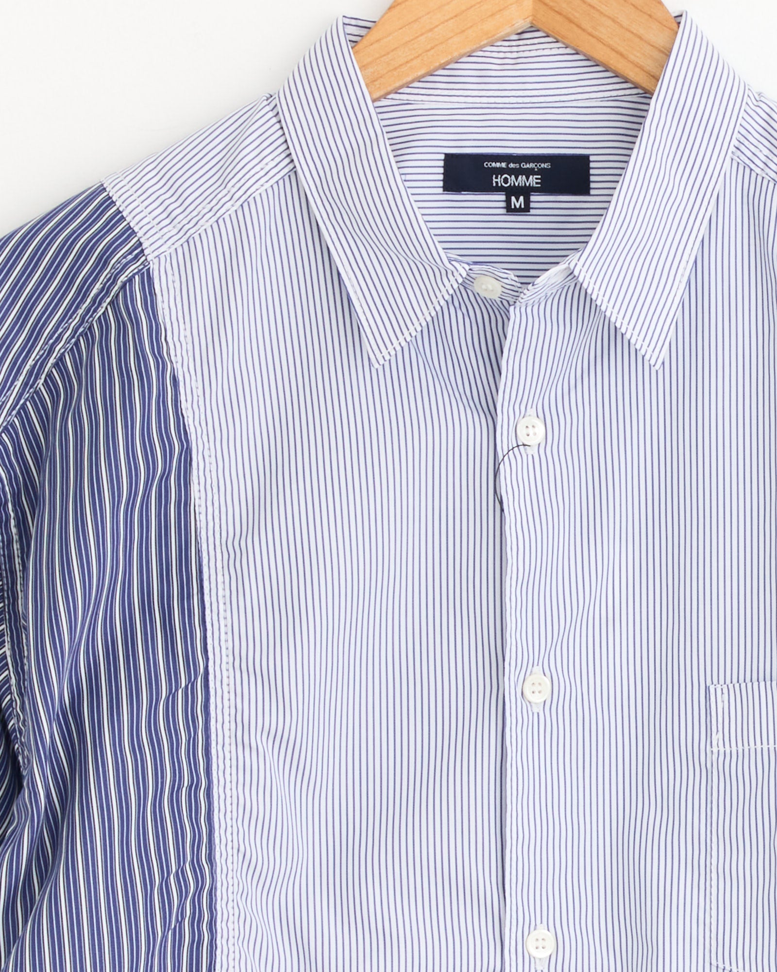 Comme Des Garçons Homme Striped Shirt in White/Navy features blue and white vertical stripes of varying widths, a pointed collar, and a right chest pocket. Displayed on a wooden hanger. Made in Japan.