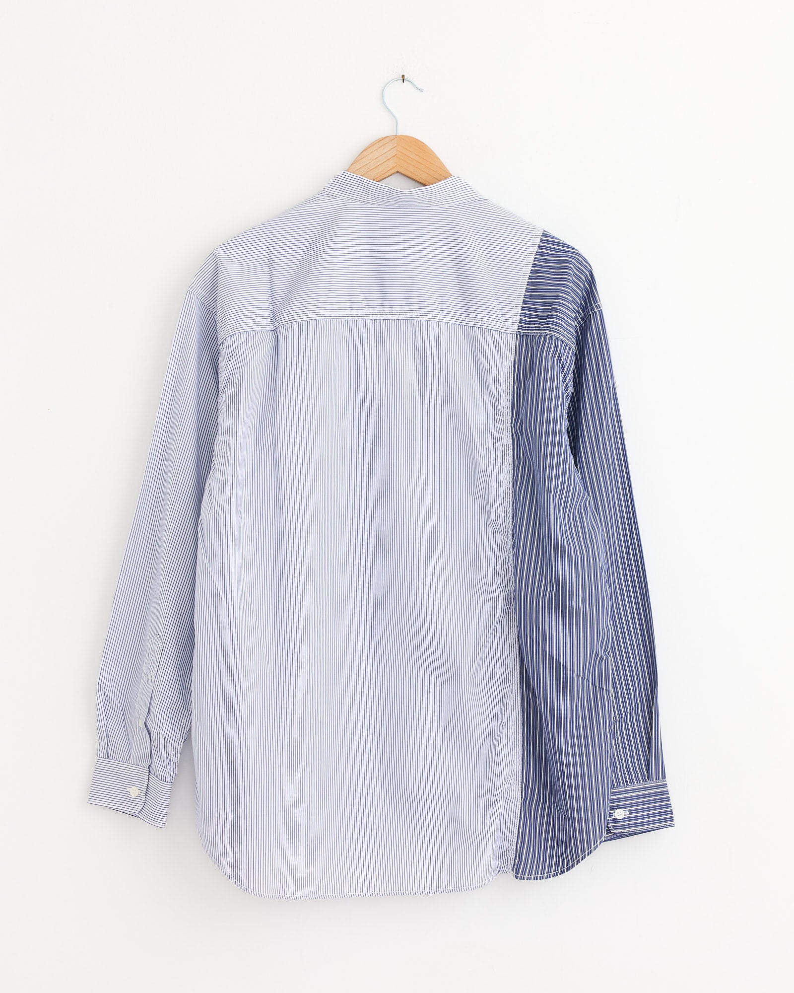 The Comme Des Garçons Homme Striped Shirt in White/Navy features mismatched vertical stripes—darker blue on one sleeve and back, lighter blue elsewhere—hung on a wooden hanger against a white wall. Made in Japan.