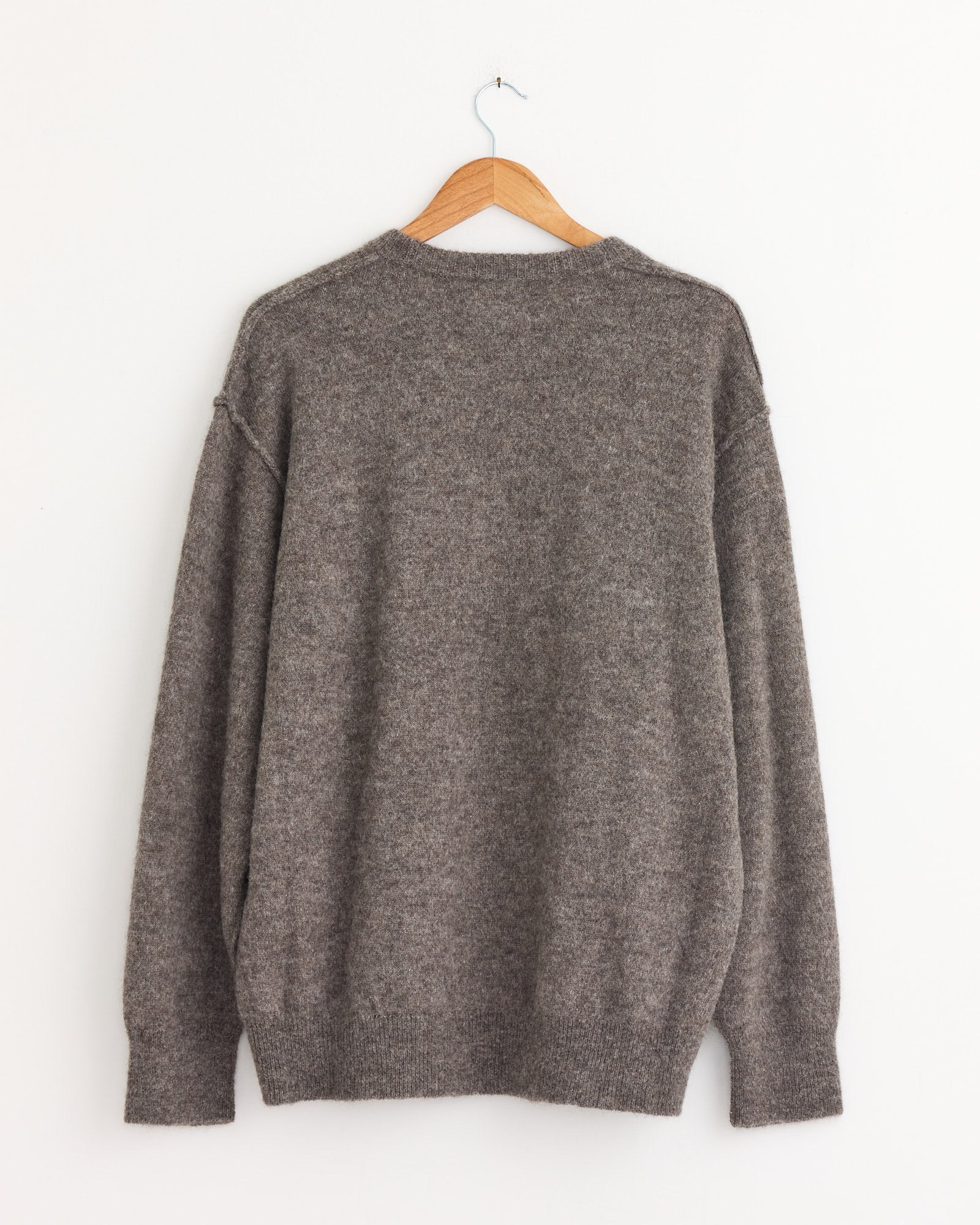 The ssstein 1294 Sweater in Greige is displayed on a wooden hanger against a white wall, shown from the back with long sleeves and ribbed collar, hem, and cuffs.