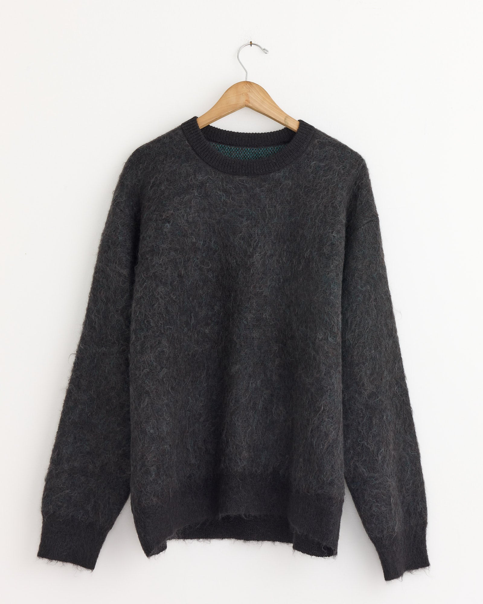 The ssstein 1305 Sweater in Mix, a fluffy dark gray kid mohair, long-sleeve style, is displayed on a wooden hanger against a plain white wall.