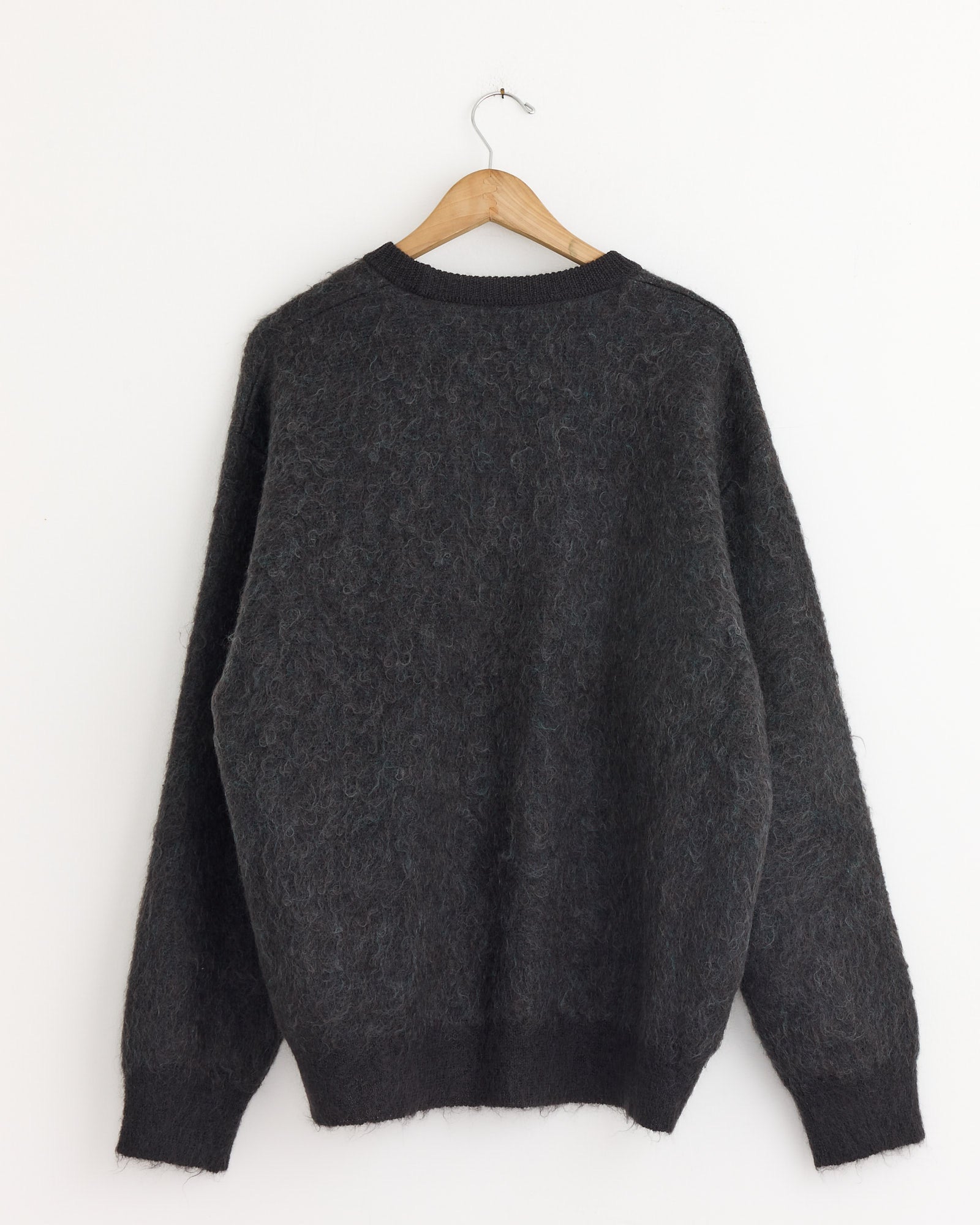 The ssstein 1305 Sweater in Mix, a dark gray fuzzy jacquard-knit style, hangs on a wooden hanger against a plain white wall, displayed from the back.