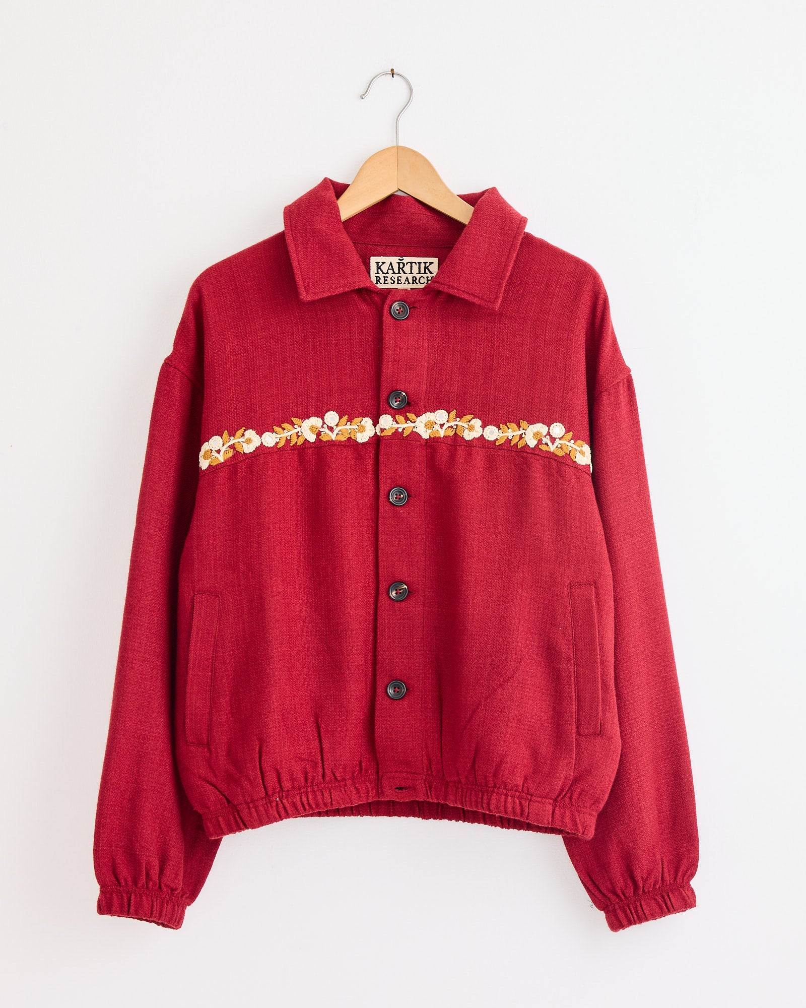 The Kartik Research Rishikesh Jacket in Red/Gold/Ecru features long sleeves, front pockets, a collar, and a chest band of embroidered yellow and white flowers. For sizing or fit questions, please use our online chat.