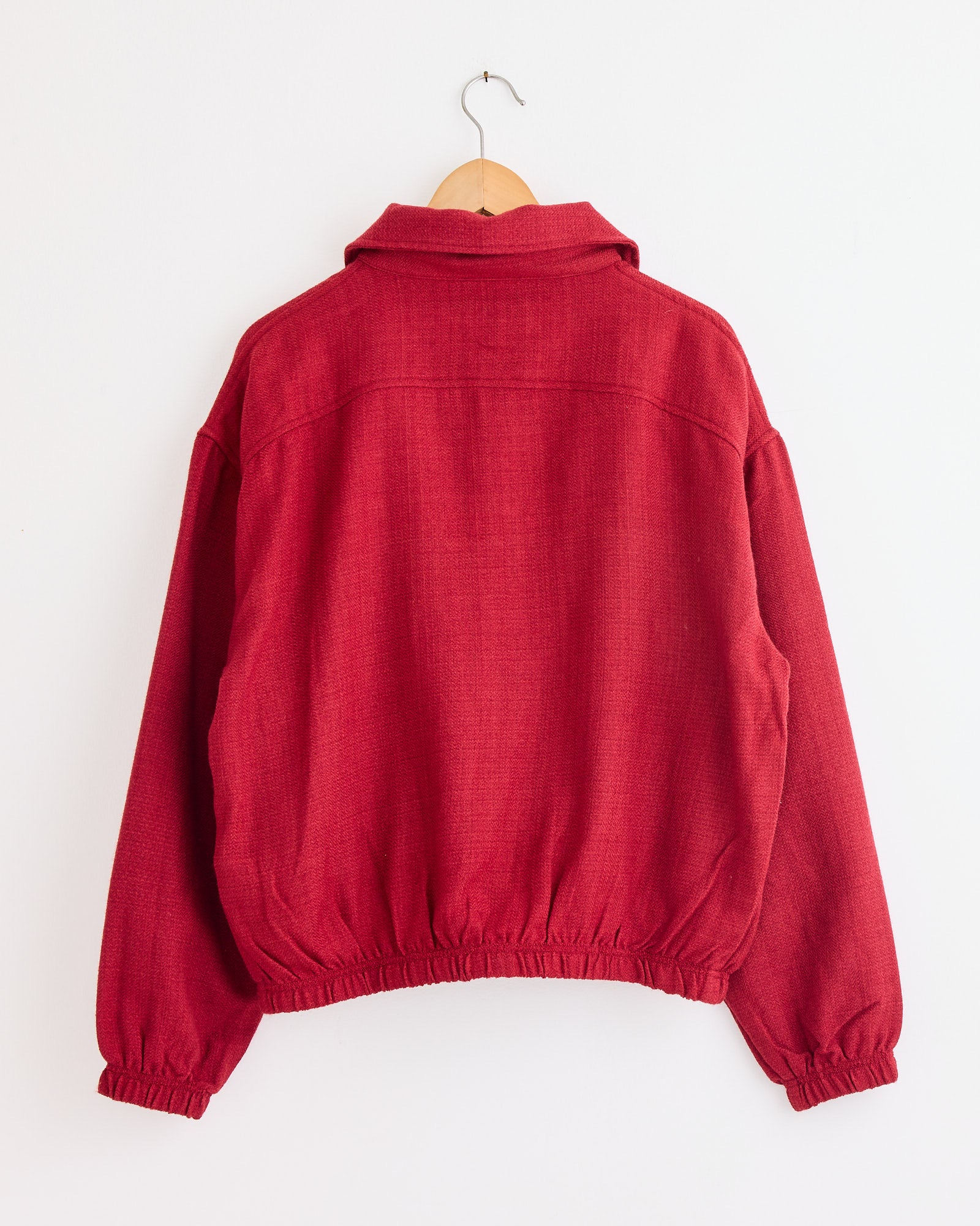 The Rishikesh Jacket in Red/Gold/Ecru by Kartik Research features long sleeves, a collar, and elastic cuffs and hem, shown from the back on a wooden hanger against a white background. Chat online for sizing or fit questions.