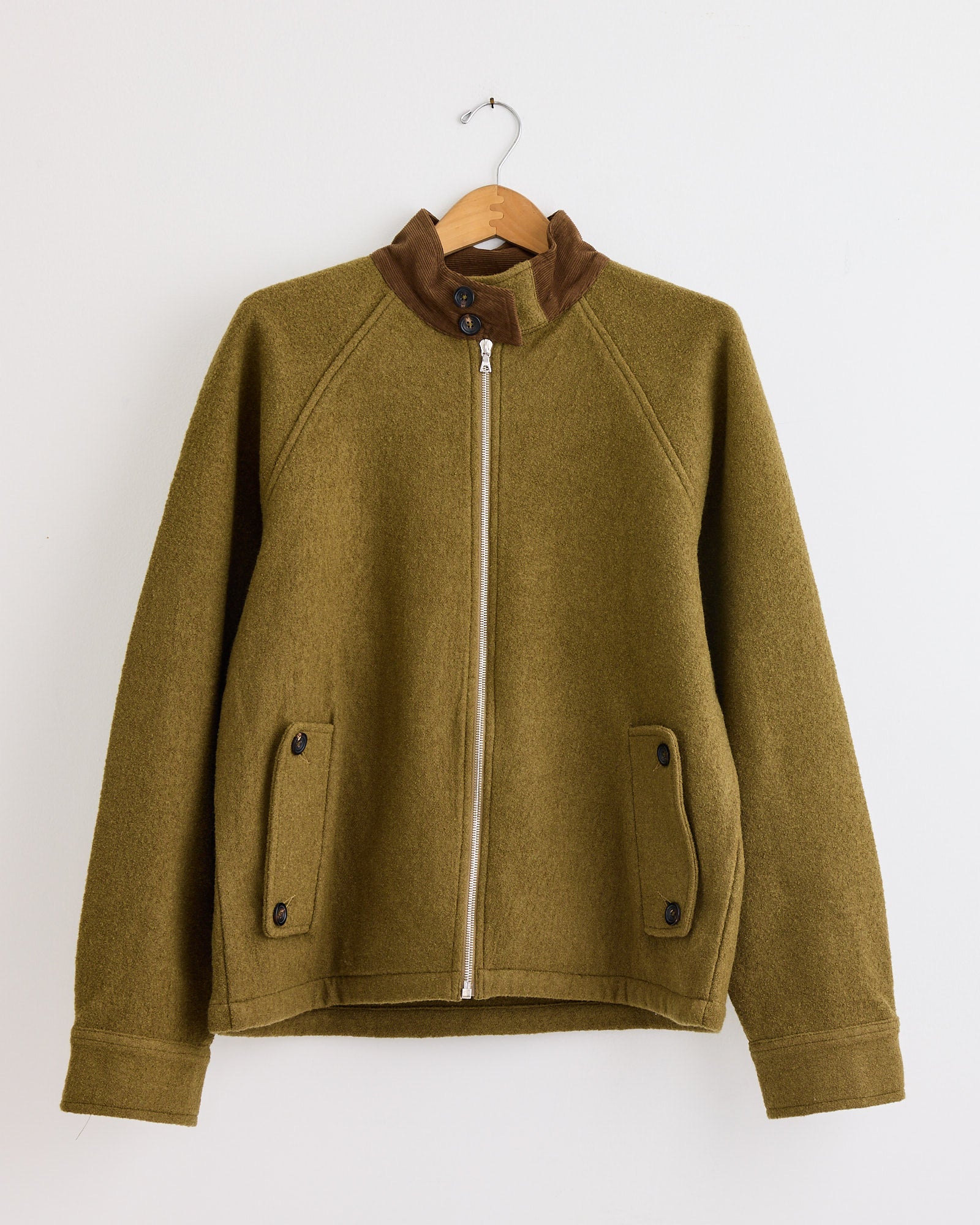 The Harrington Jacket in Moss/Teak by Kartik Research, crafted from handwoven merino wool by Indian artisans, features a front zipper, brown collar detail, and two buttoned pockets, shown on a wooden hanger against a white wall.