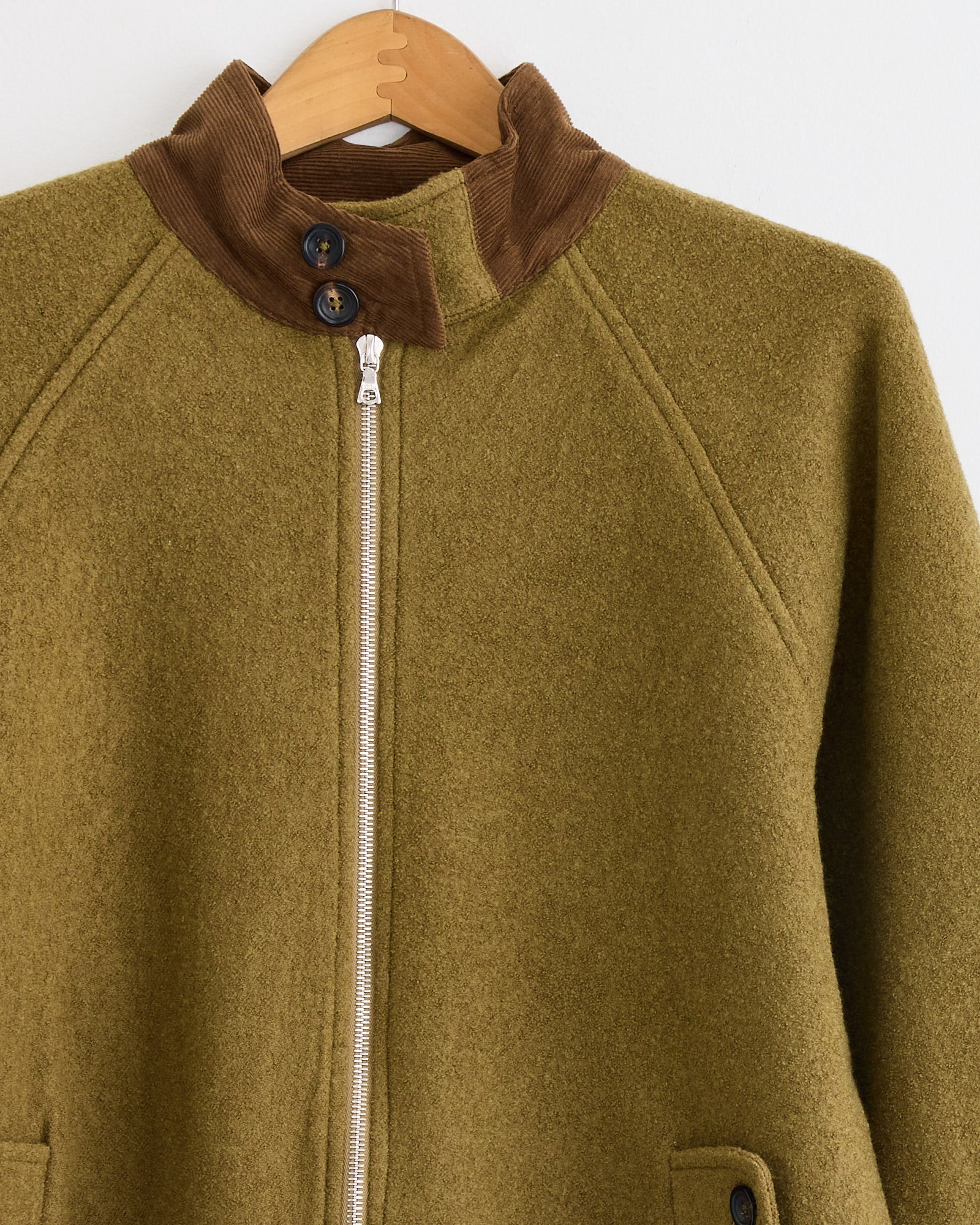 The Kartik Research Harrington Jacket in Moss/Teak features handwoven merino wool by Indian artisans, a brown corduroy collar, black neck buttons, silver zipper, and front pockets—shown on a wooden hanger against a white background.