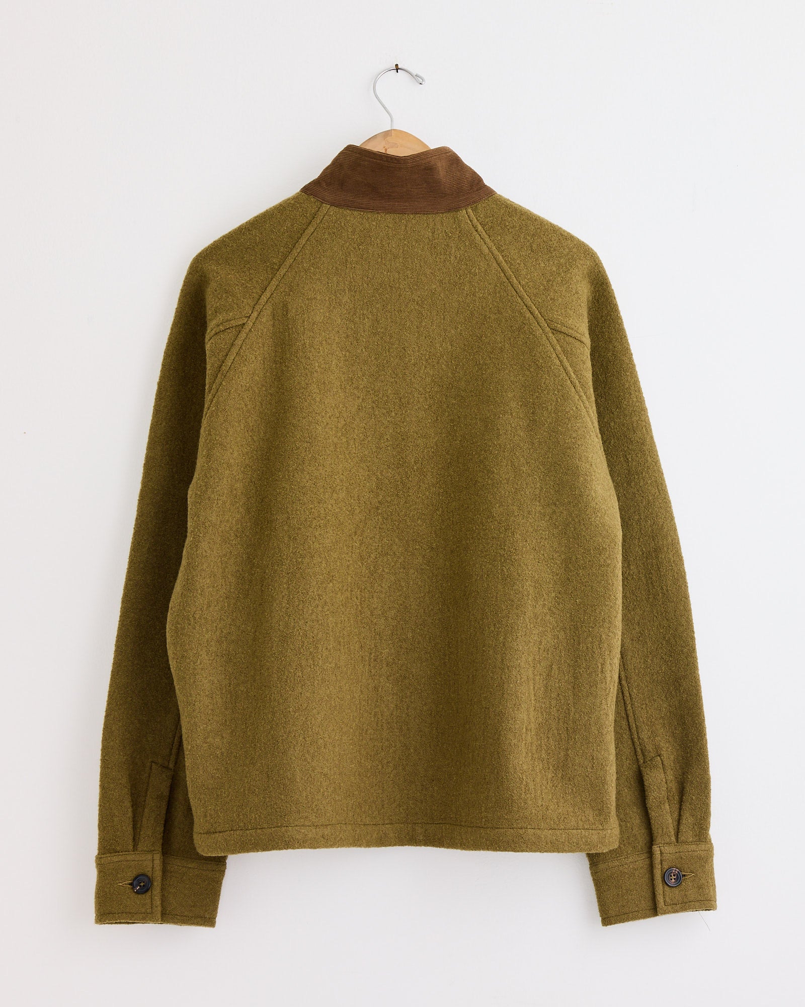 The Kartik Research Harrington Jacket in Moss/Teak, handwoven from merino wool by Indian artisans, has a brown suede collar, buttoned cuffs, and clean lines, shown from the back on a wooden hanger against a white wall.