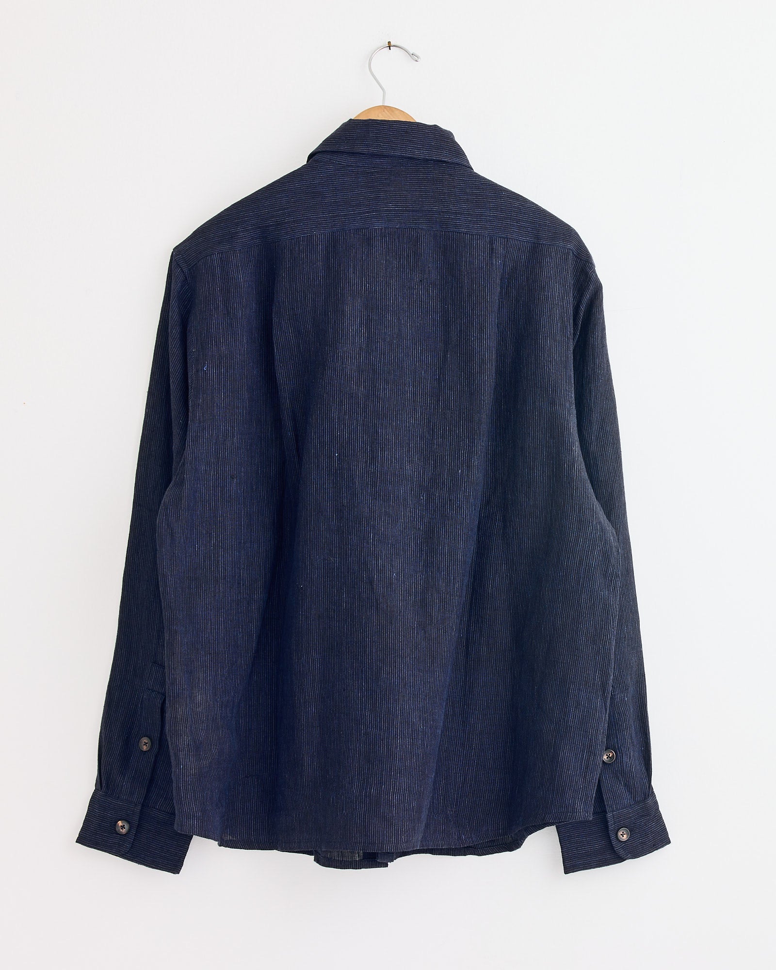 The Double Layer Shirt in Indigo/Taupe by Kartik Research is a hand-embroidered, long-sleeve linen shirt shown from the back, hanging on a wooden hanger against a plain white wall.