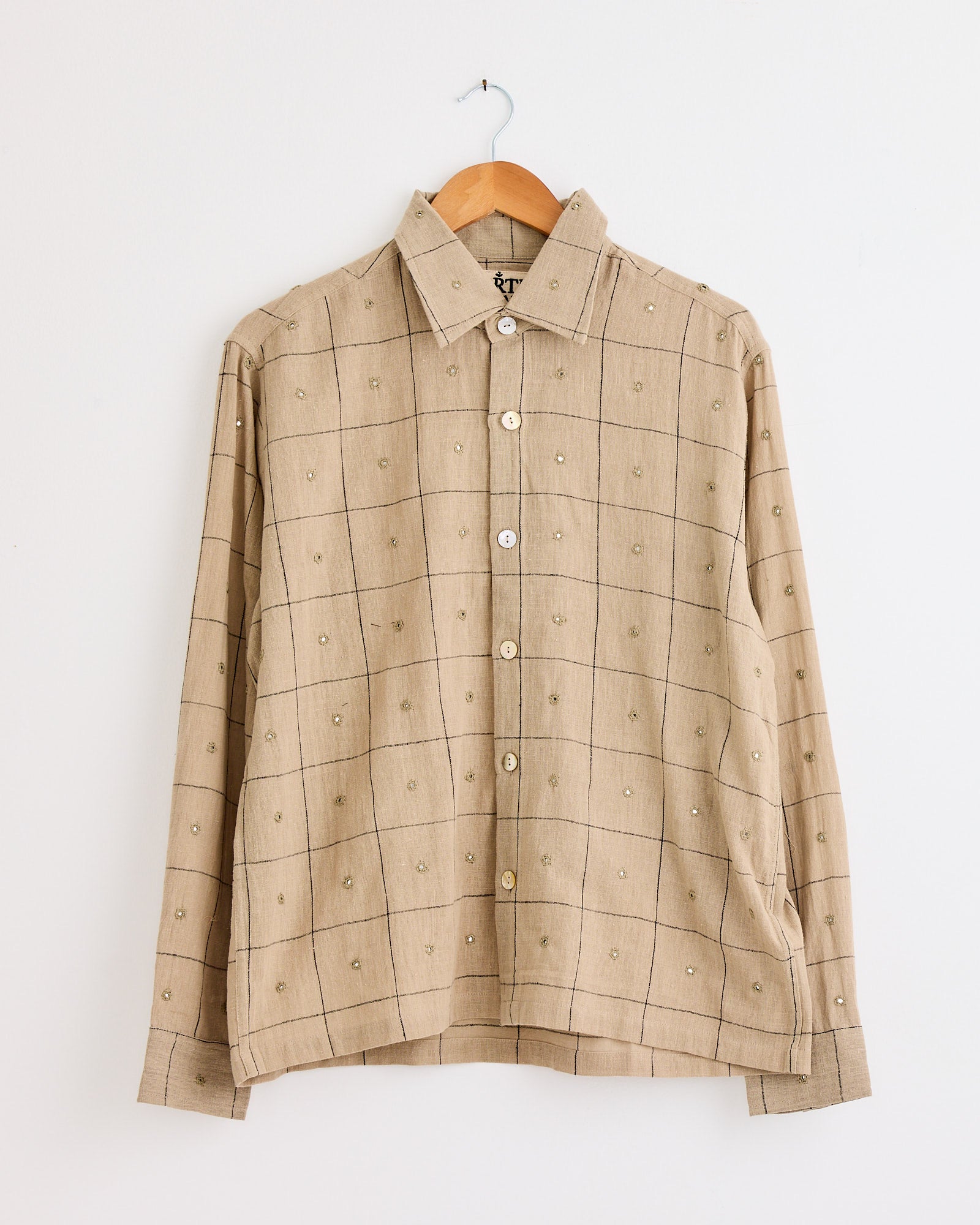 The Kartik Research Hand Embroidered Mirror Box Shirt in Beige/Indigo features long sleeves, a grid pattern with small dot details, and intricate hand embroidery. Displayed on a wooden hanger against a plain white wall.