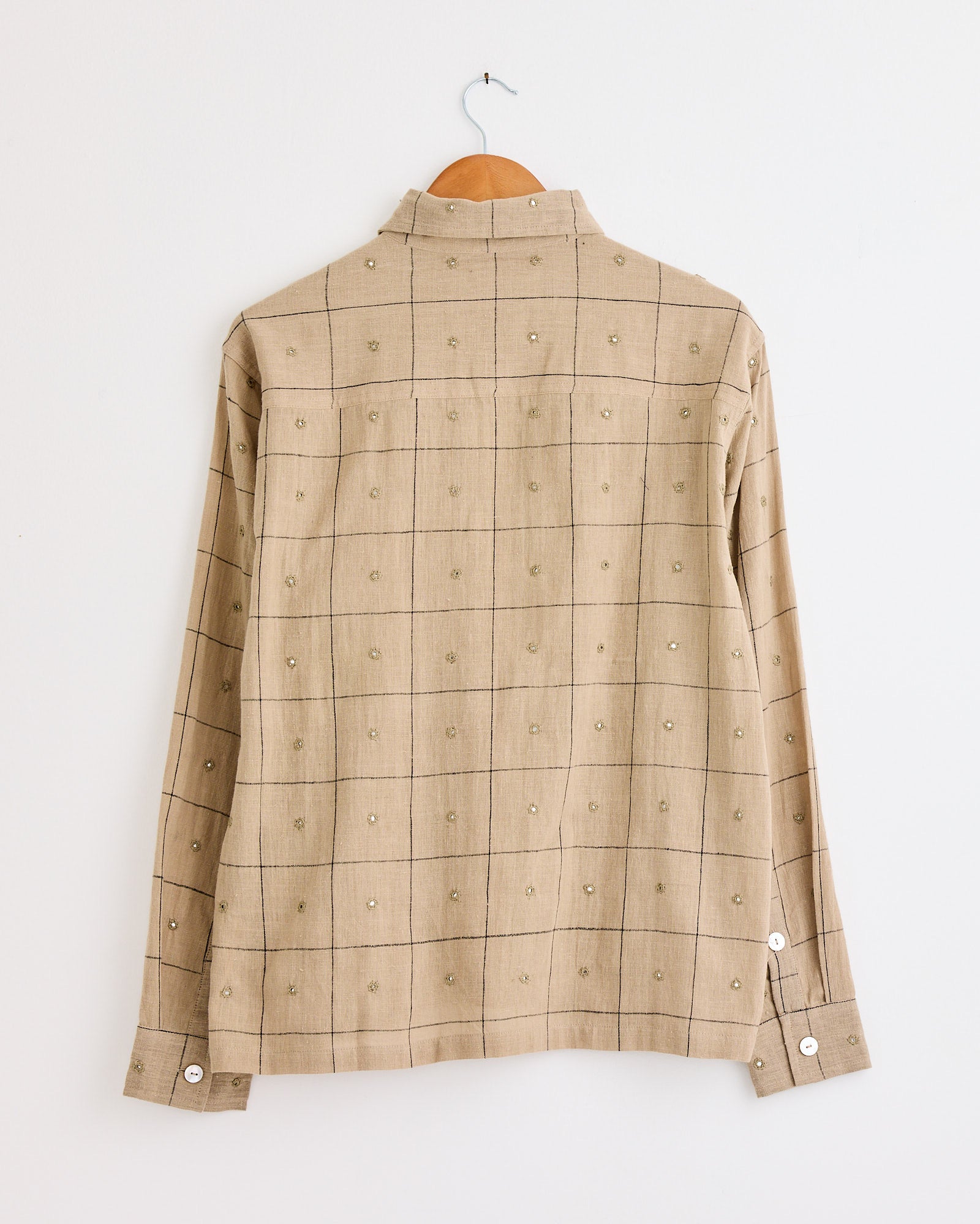 A beige Hand Embroidered Mirror Box Shirt by Kartik Research hangs on a wooden hanger, showcasing a grid pattern with embroidered details and white cuff buttons, displayed from the back against a white wall.